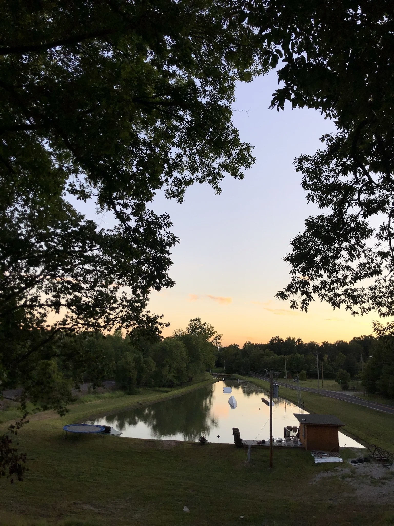 A calm canal reflecting the sky at sunset, framed by trees and a small wooden structure on the right, with a trampoline on the left and boats docked along the water.