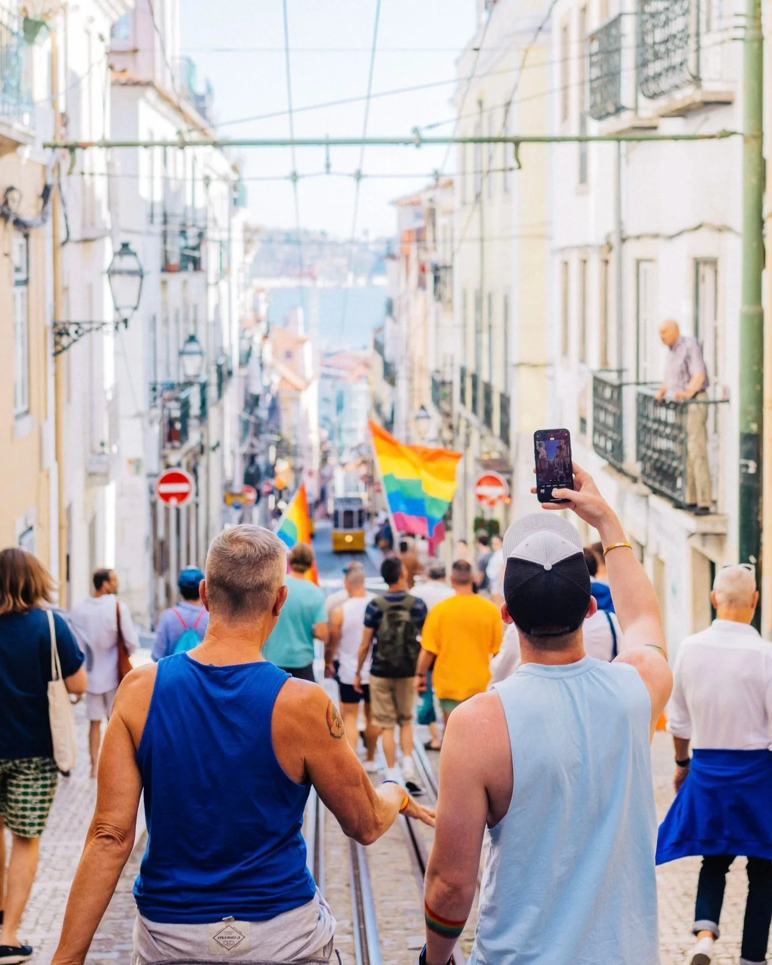 LGBTQ+ pride parade marching down a narrow cobblestone street in Lisbon, Portugal, with a rainbow flag ahead.