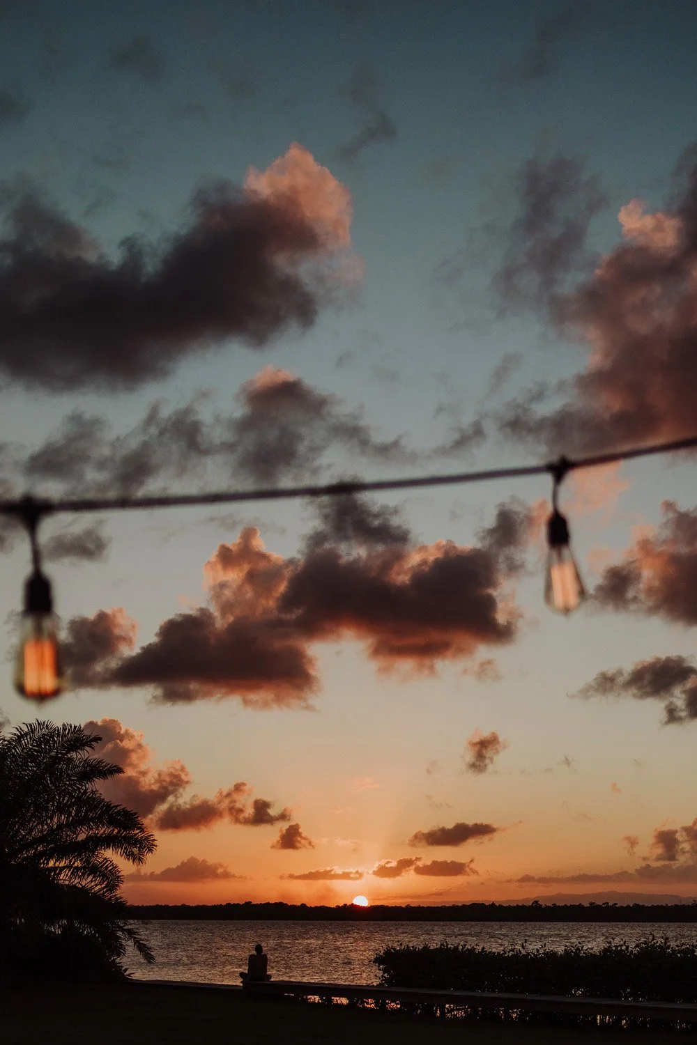 Sunset over a body of water with dark clouds, a person sitting on the shore, and string lights hanging in the foreground.
