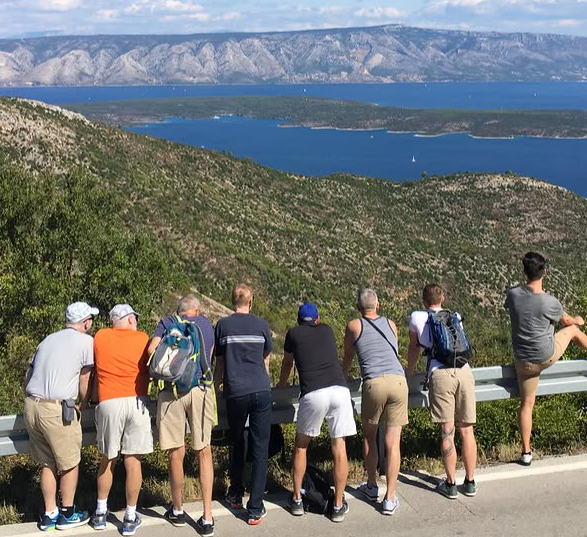 Group of travelers lined up at a roadside barrier overlooking a coastal viewpoint, gazing at hills, islands, and sea in the distance.
