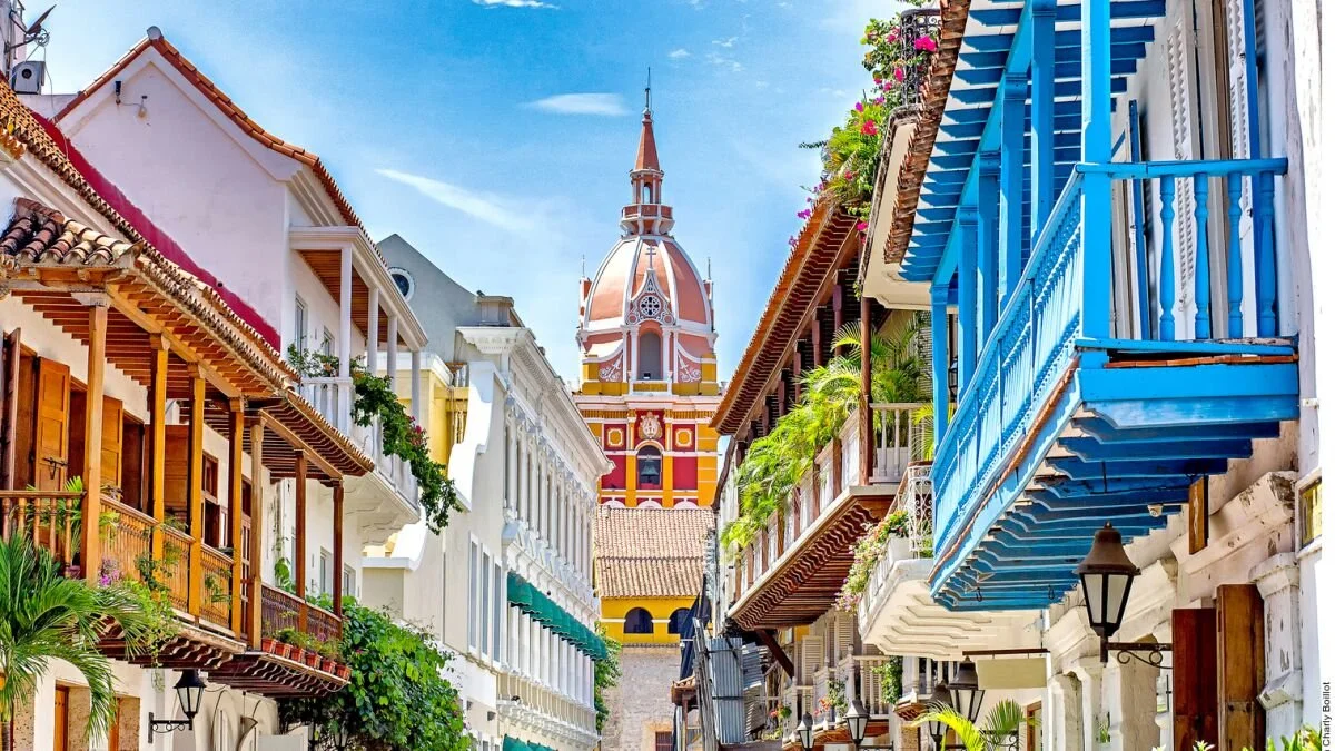 Colorful European-style street with white buildings, wooden balconies, and a tall church tower in the background under a blue sky.
