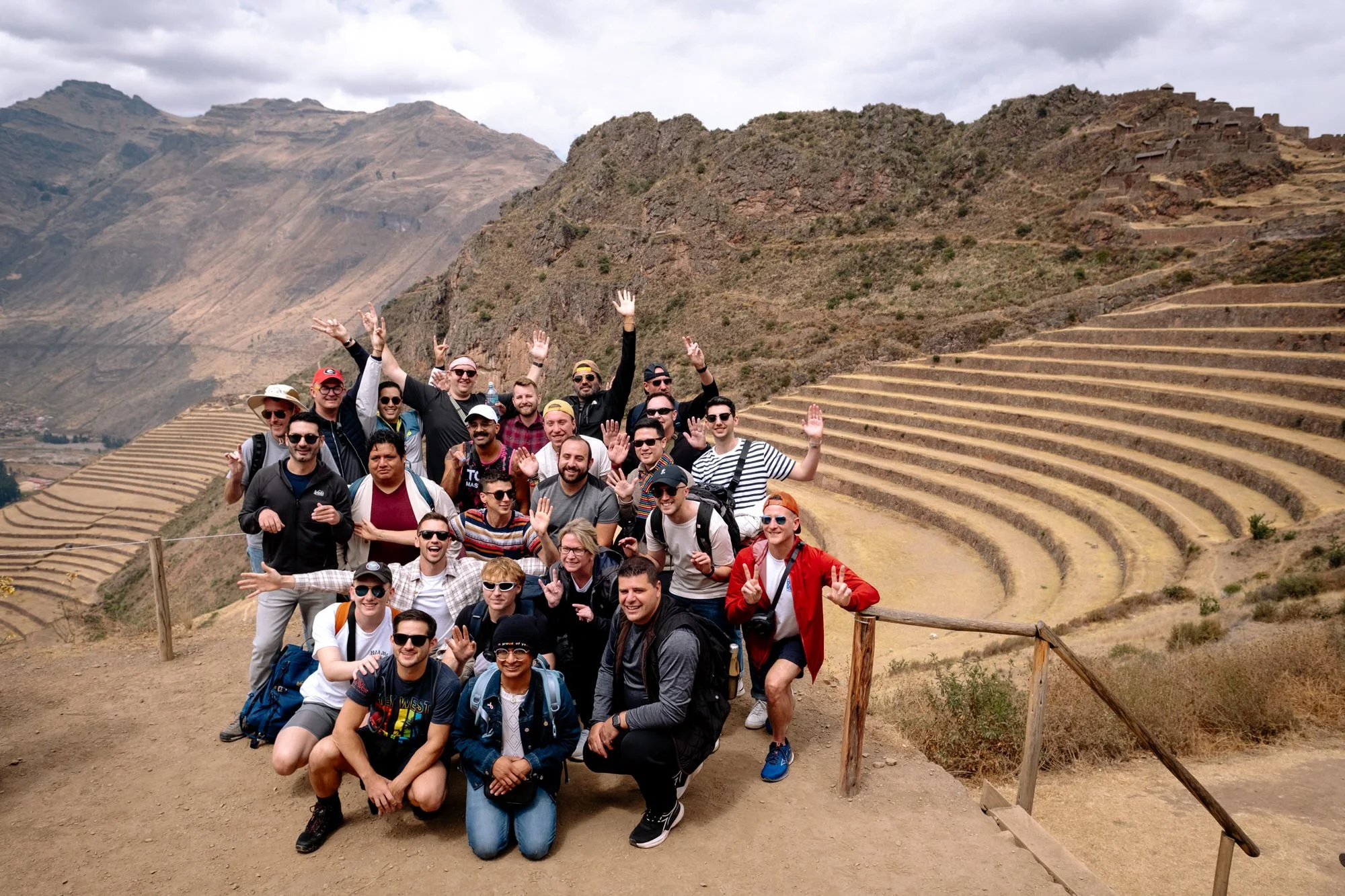 LGBTQ+ travel group posing at Pisac Inca terraces in Peru's Sacred Valley, mountains behind them.