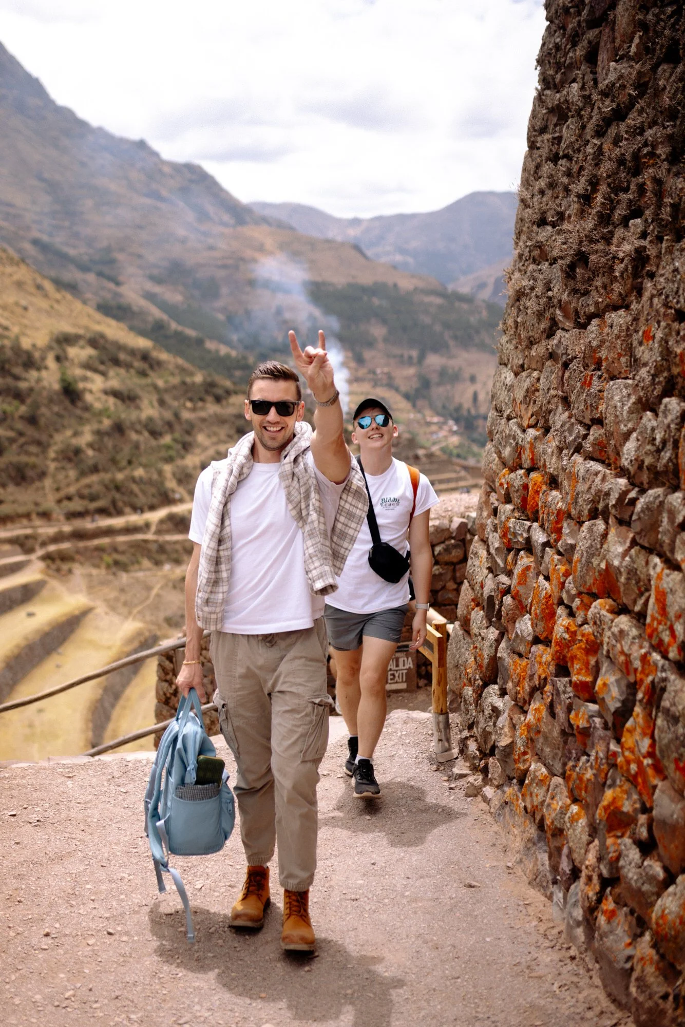 Two travelers walking the stone exit path at Pisac ruins, with Inca terraces and Sacred Valley below.