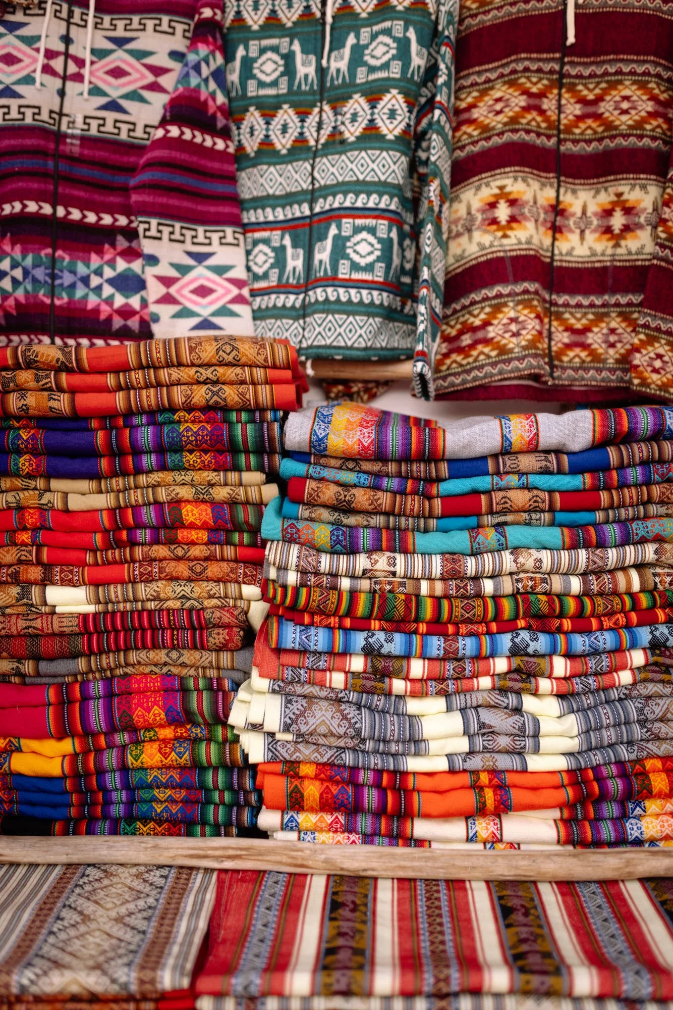 Stacks of colorful hand-woven Andean textiles with geometric patterns at a Sacred Valley market stall.