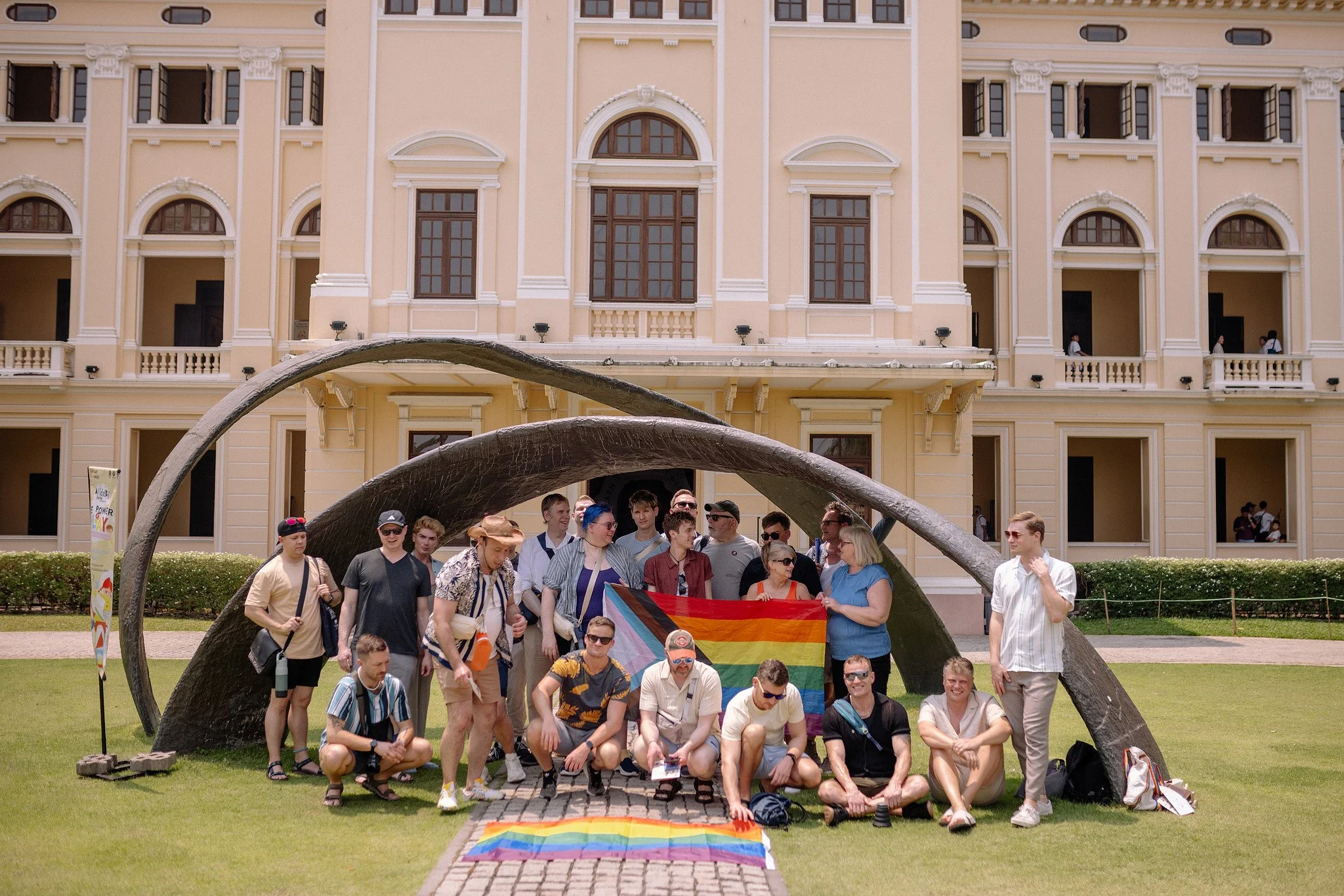  Sidequests travel group gathering under a sculptural archway with pride flags spread on the lawn 
