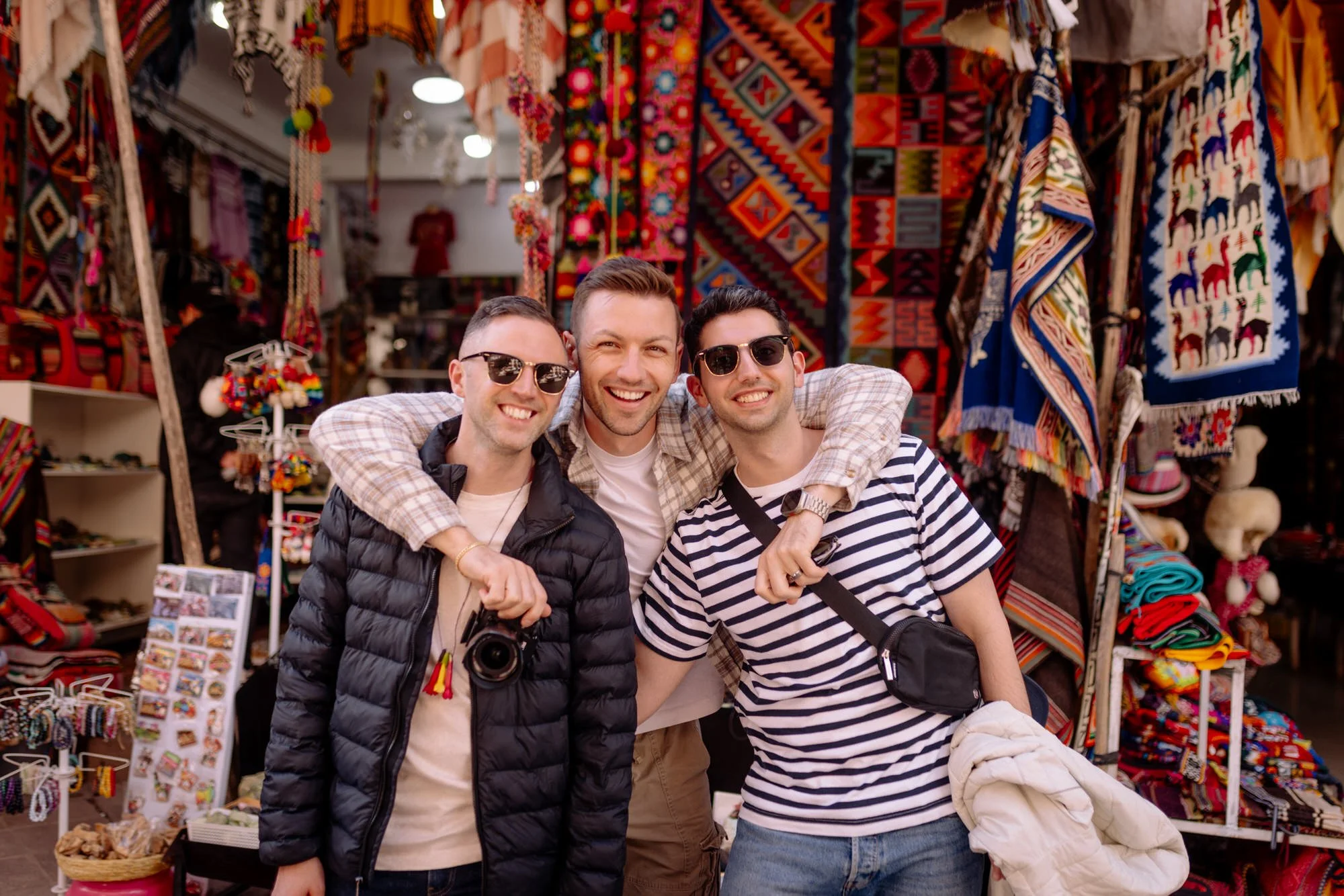 Three smiling friends posing together at a colorful textile market stall in Pisaq, Peru.