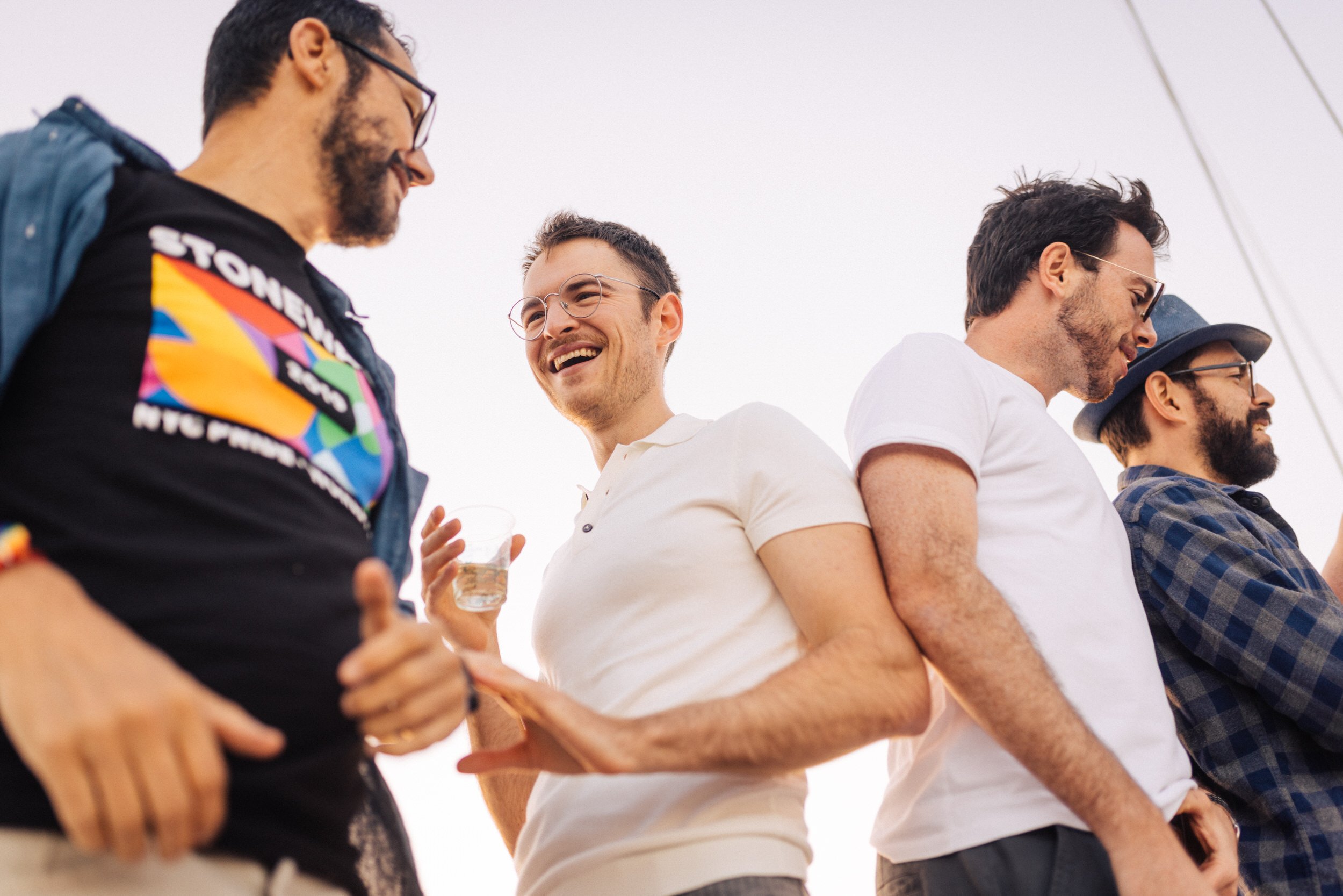 Group of men laughing at an outdoor LGBTQ+ gathering in Lisbon, one wearing a Stonewall NYC Pride shirt.