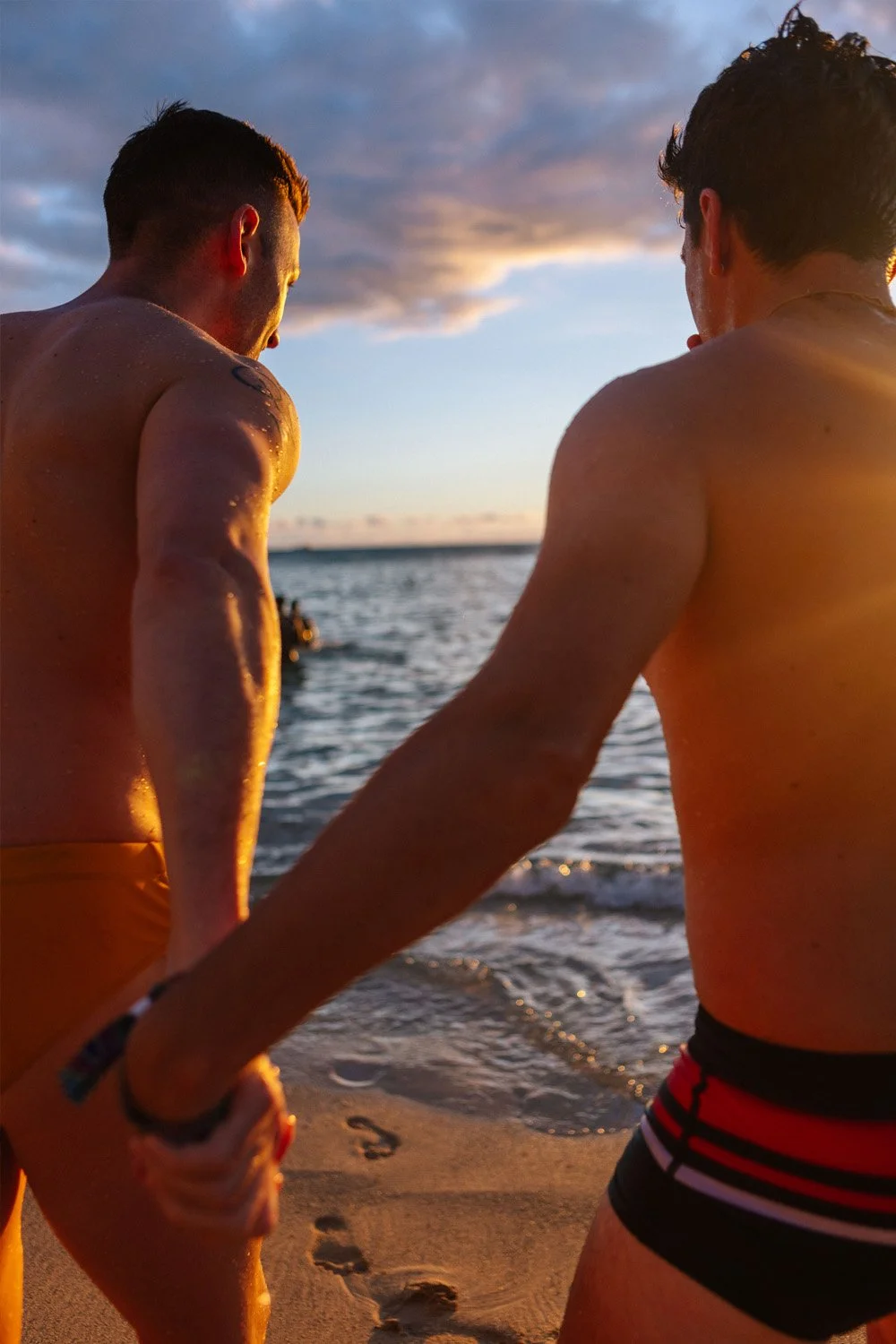 Two men holding hands and walking on the beach at sunset, near the water