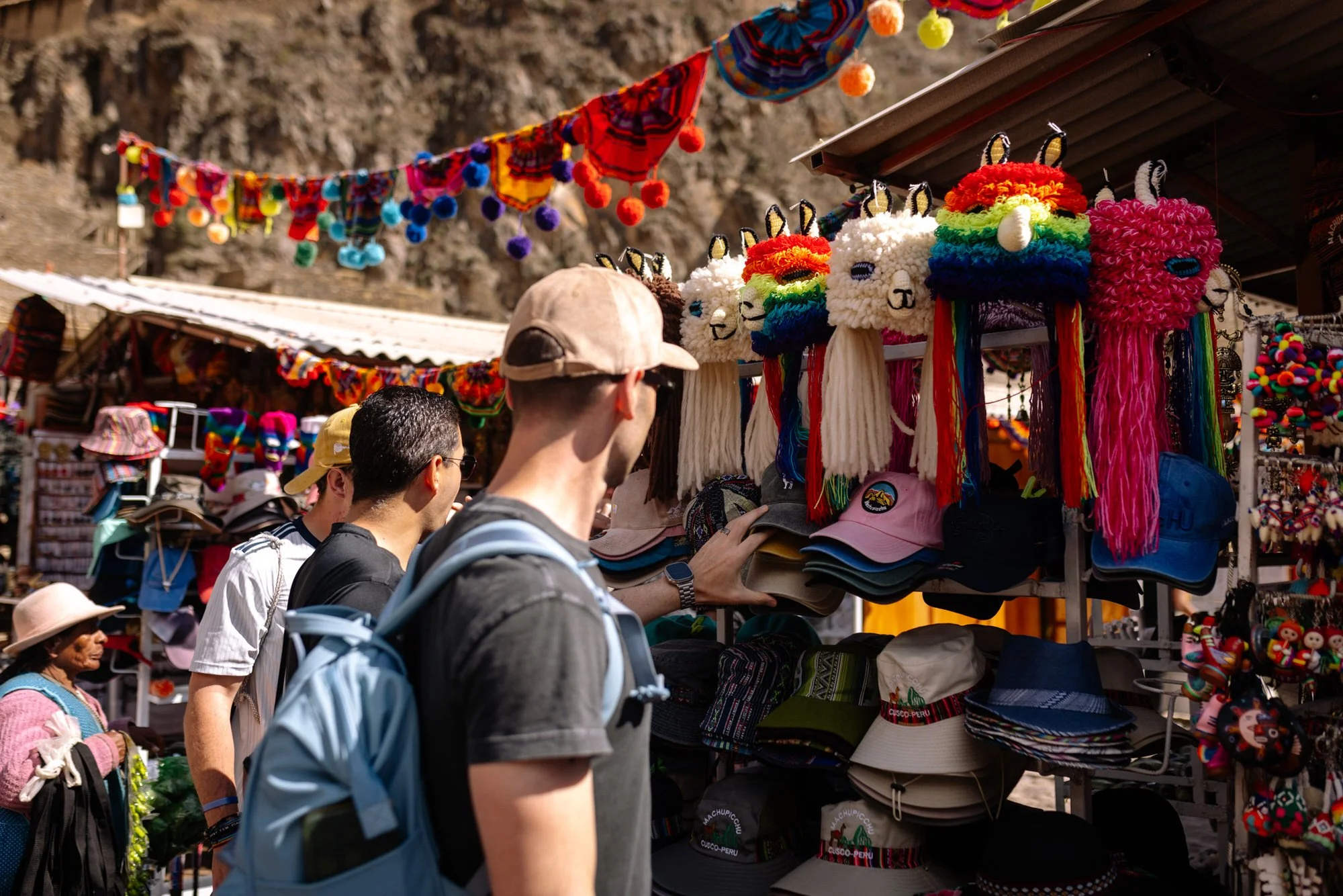 Tourists browsing colorful llama hats and caps at an open-air souvenir market in Peru's Sacred Valley.