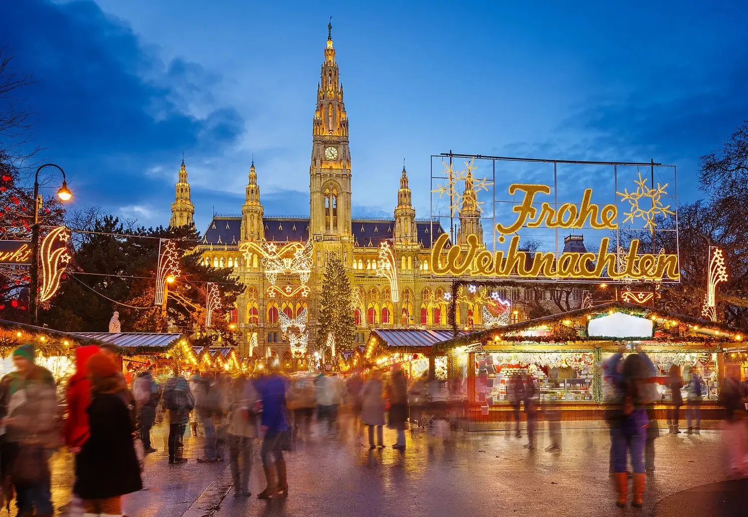 Festively decorated Christmas market with a historic building in the background and a sign saying 'Frohe Weihnachten' in German, indicating Christmas celebration.