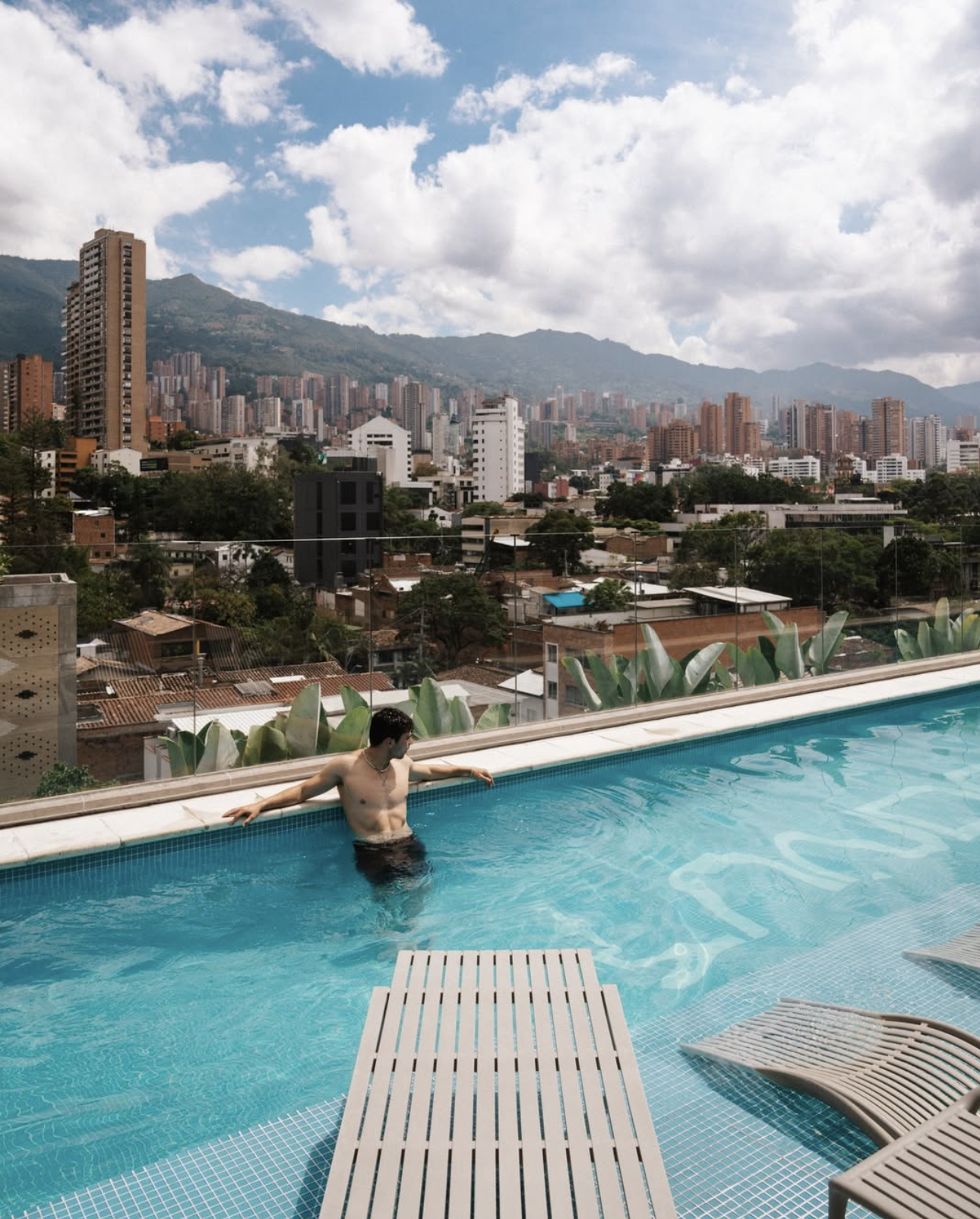 A man relaxing in a rooftop swimming pool with a city skyline and mountains in the background.