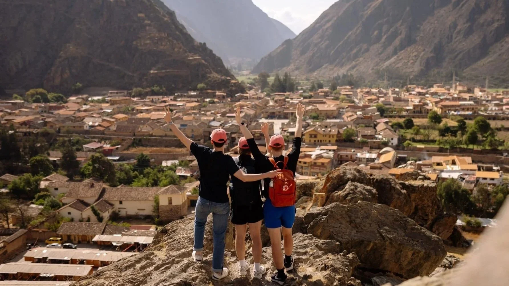 3 guys wearing pink hats throw their hands in the air while looking out over boulders at a small town in a valley