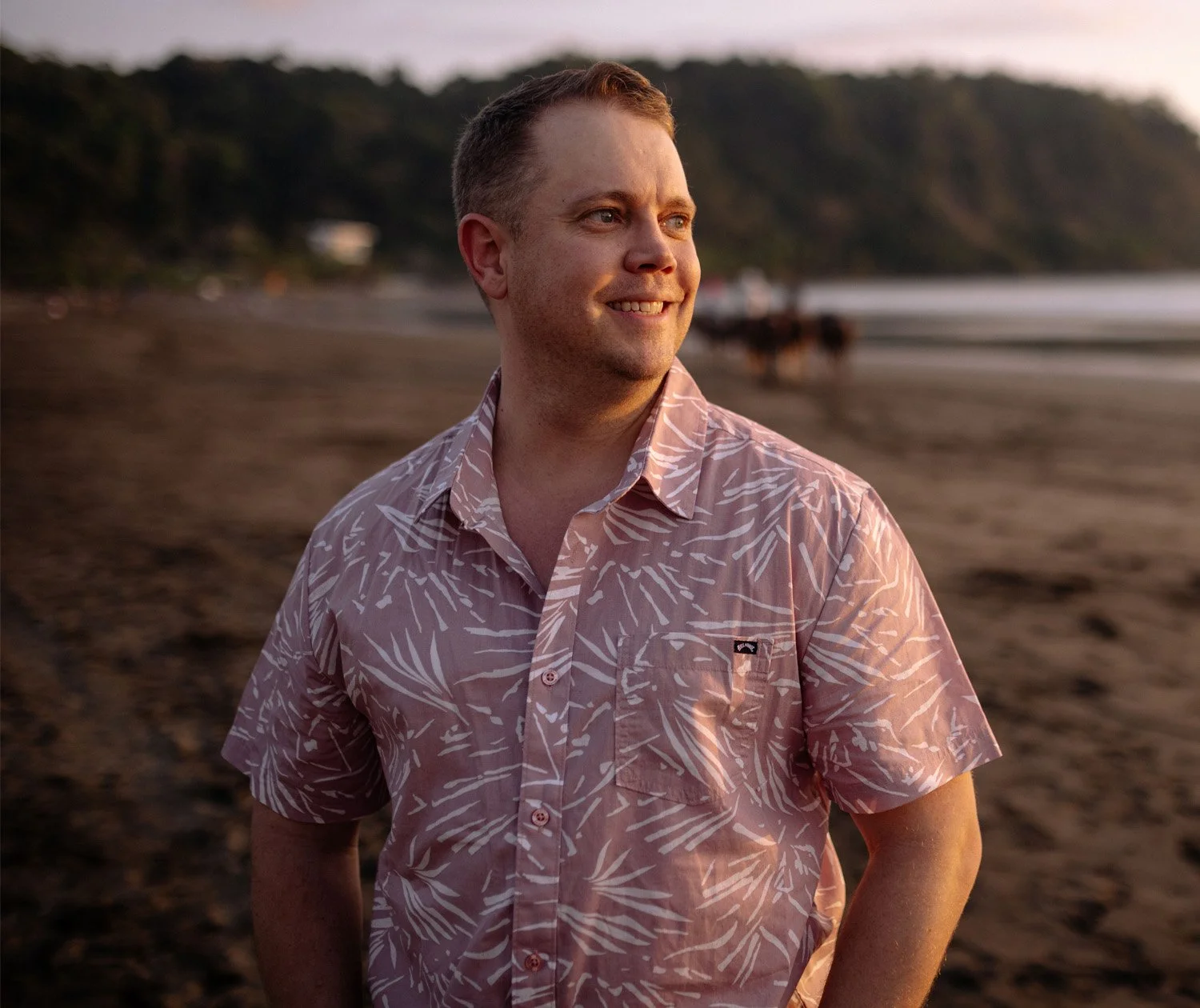 A smiling man wearing a pink tropical shirt stands on a beach at sunset with waves and hills in the background.