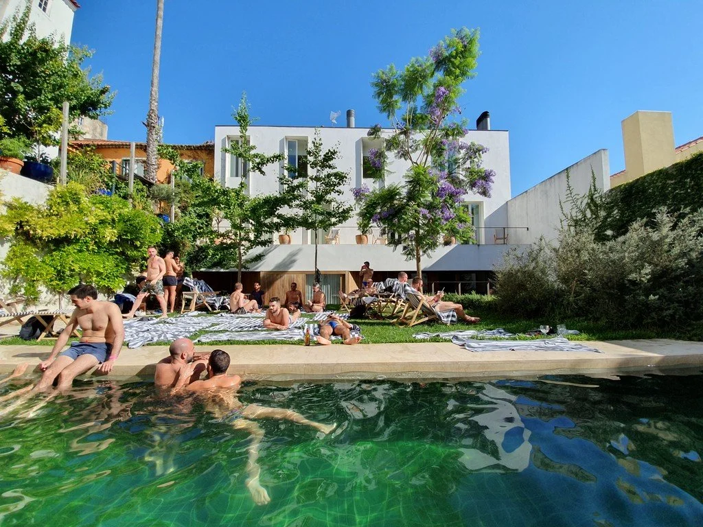men gather, sunbathe and swim in a pool together in a courtyard in front of trees and a white building