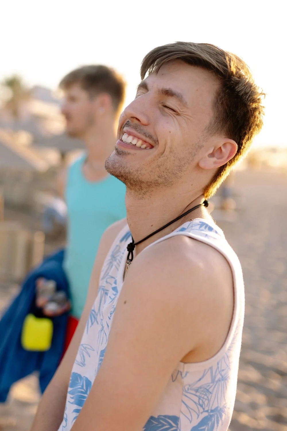 Smiling young man with short brown hair and light facial hair, wearing a sleeveless shirt with blue leaf patterns and a black choker, standing on the beach during sunset, with another young man in a blue shirt blurred in the background.