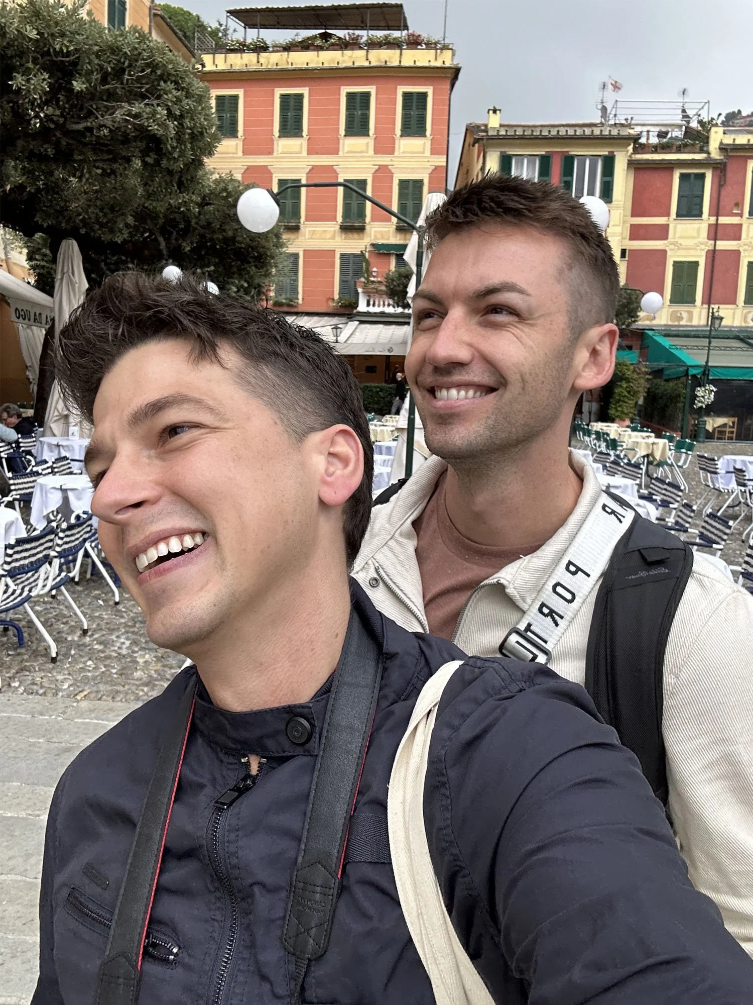 Two smiling young men taking a selfie outdoors in a city square with colorful buildings, outdoor dining tables, and decorative street lamps behind them.