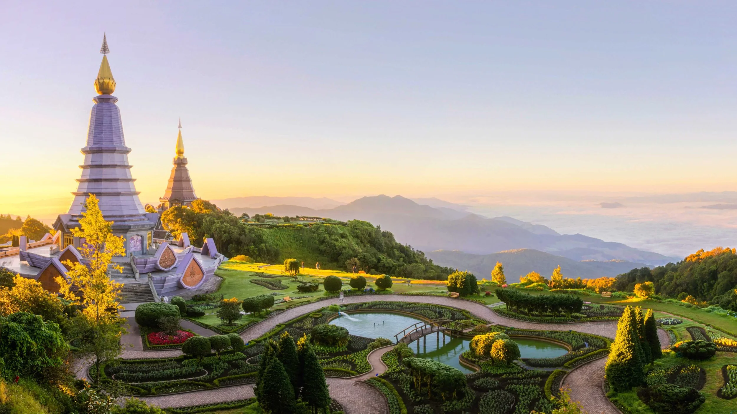 Scenic view of a garden with white and gold pagoda-style structures, a pond, and lush green trees, with mountains and a sky at sunrise or sunset in the background.