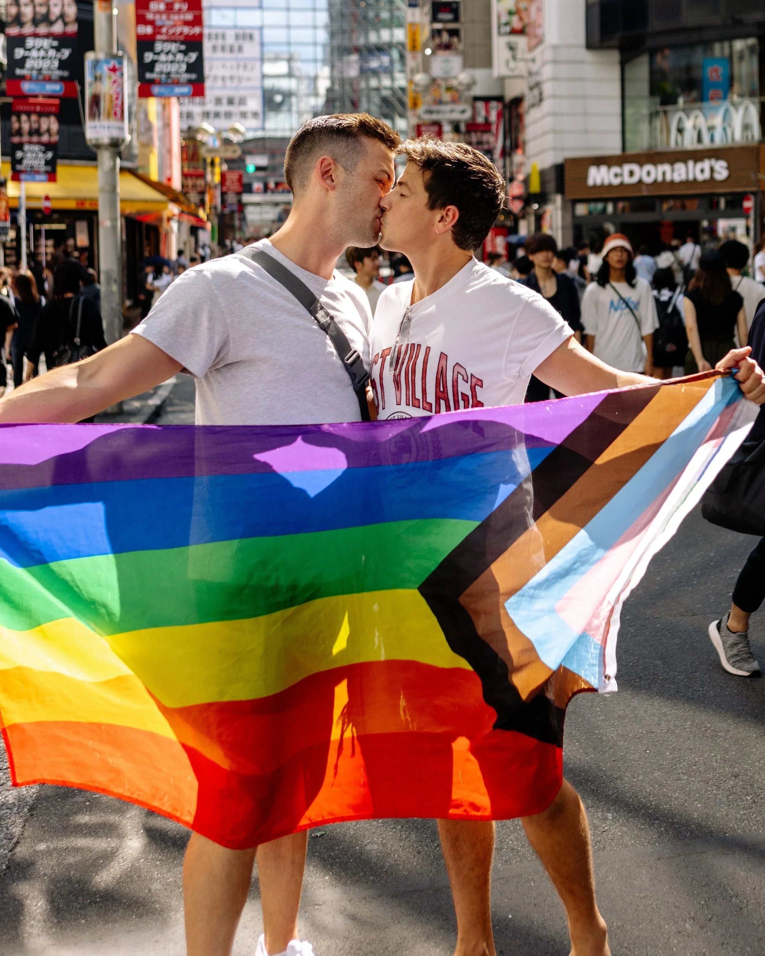 A gay couple kissing in busy pedestrian street holding a rainbow flag