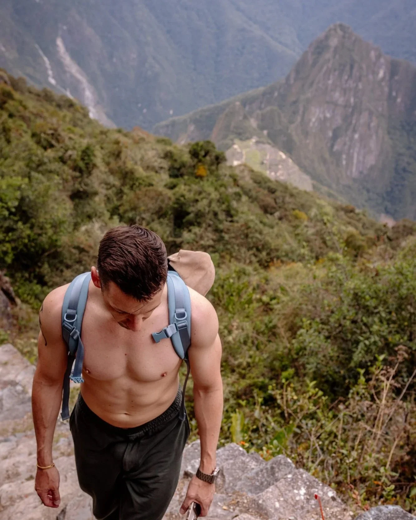 Shirtless hiker climbing stone steps on Huayna Picchu with Machu Picchu ruins visible below.