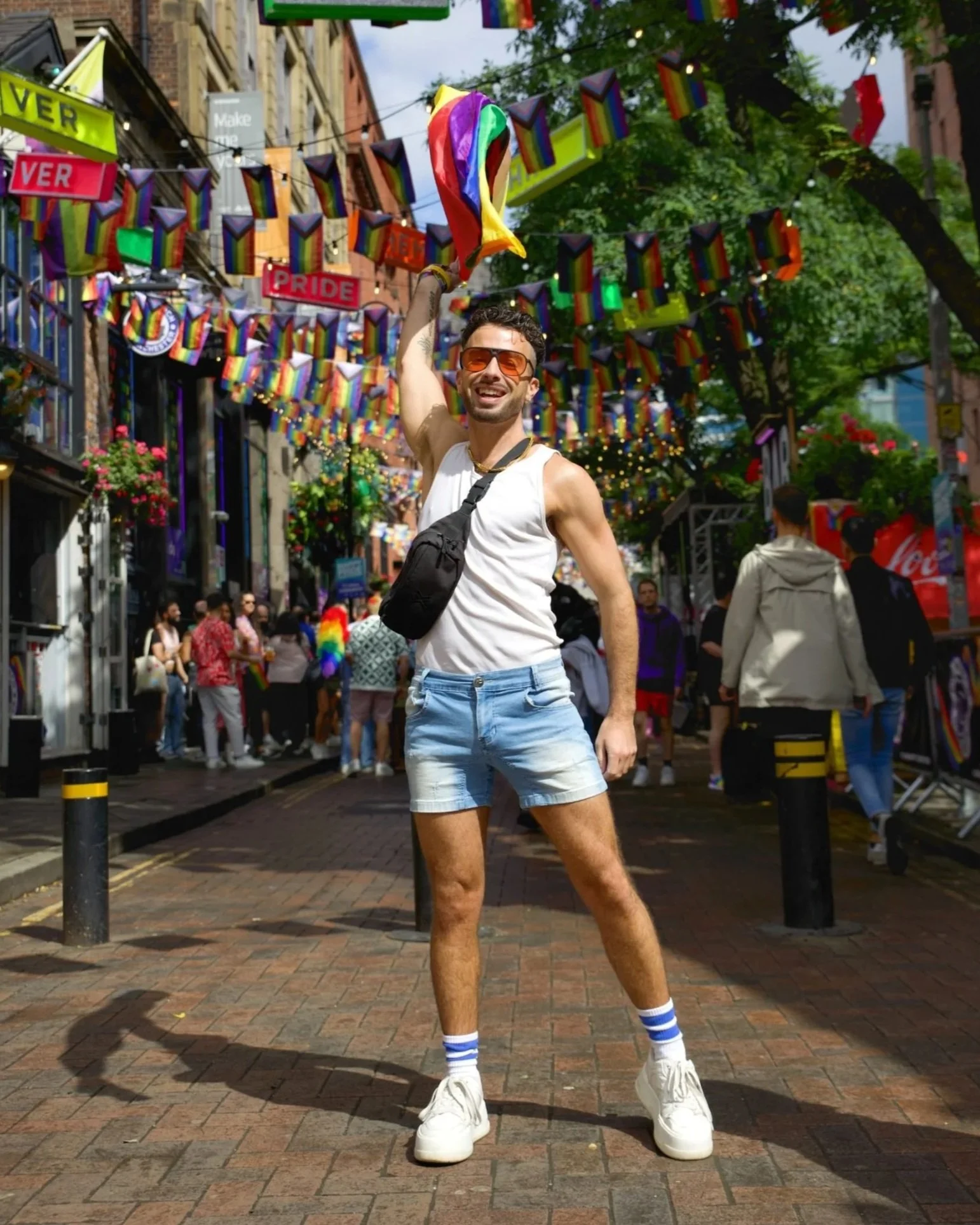 a man in a white tank-top waves a rainbow flag in front of pride flag banners down a brick street