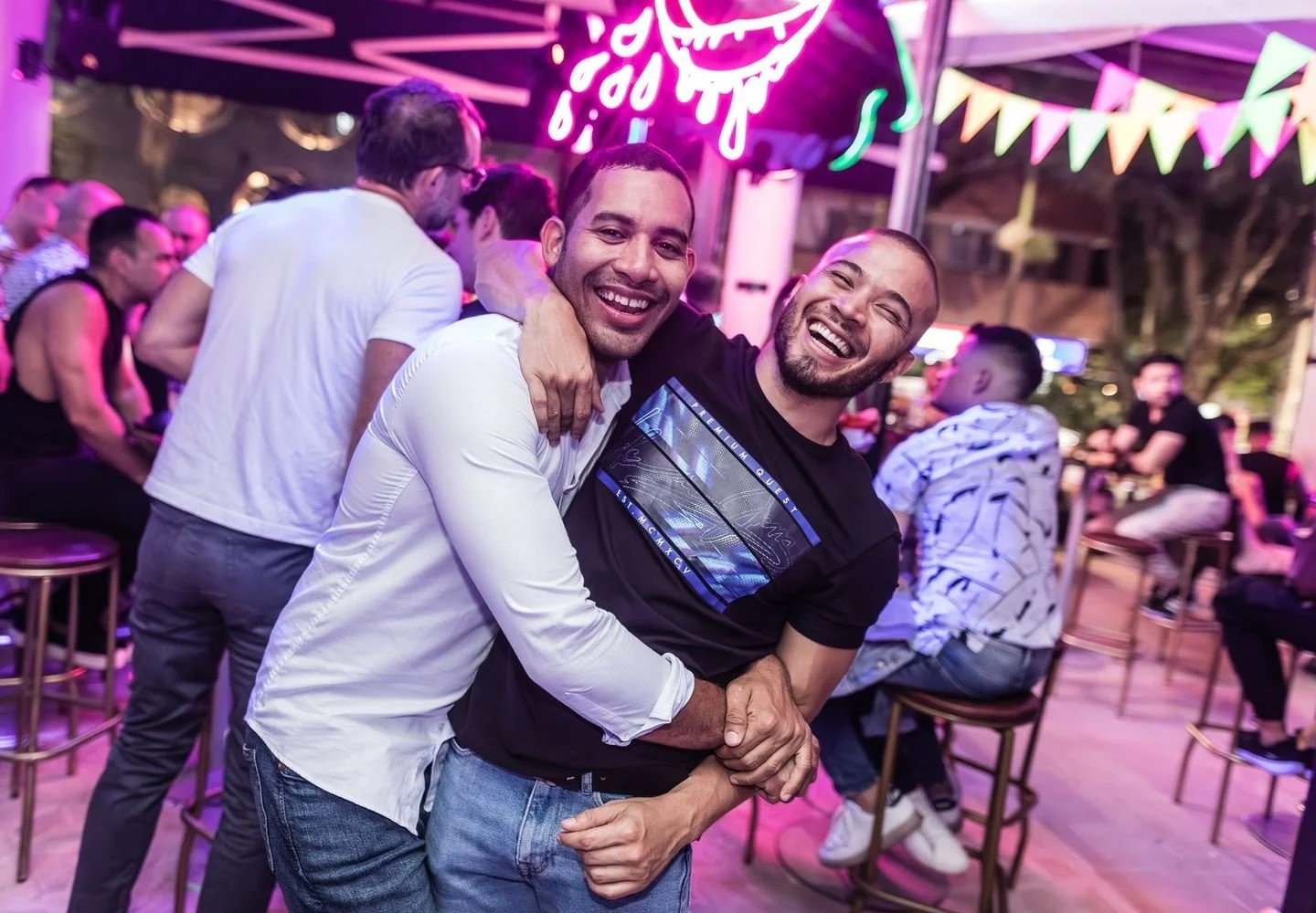 Two men laughing and embracing at a vibrant neon-lit bar, celebrating nightlife in Colombia.