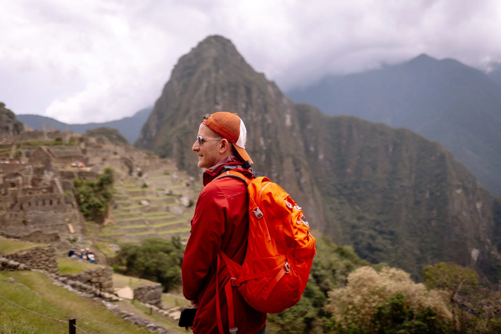 Hiker in red jacket and orange pack overlooking Machu Picchu ruins and Huayna Picchu mountain, Peru.