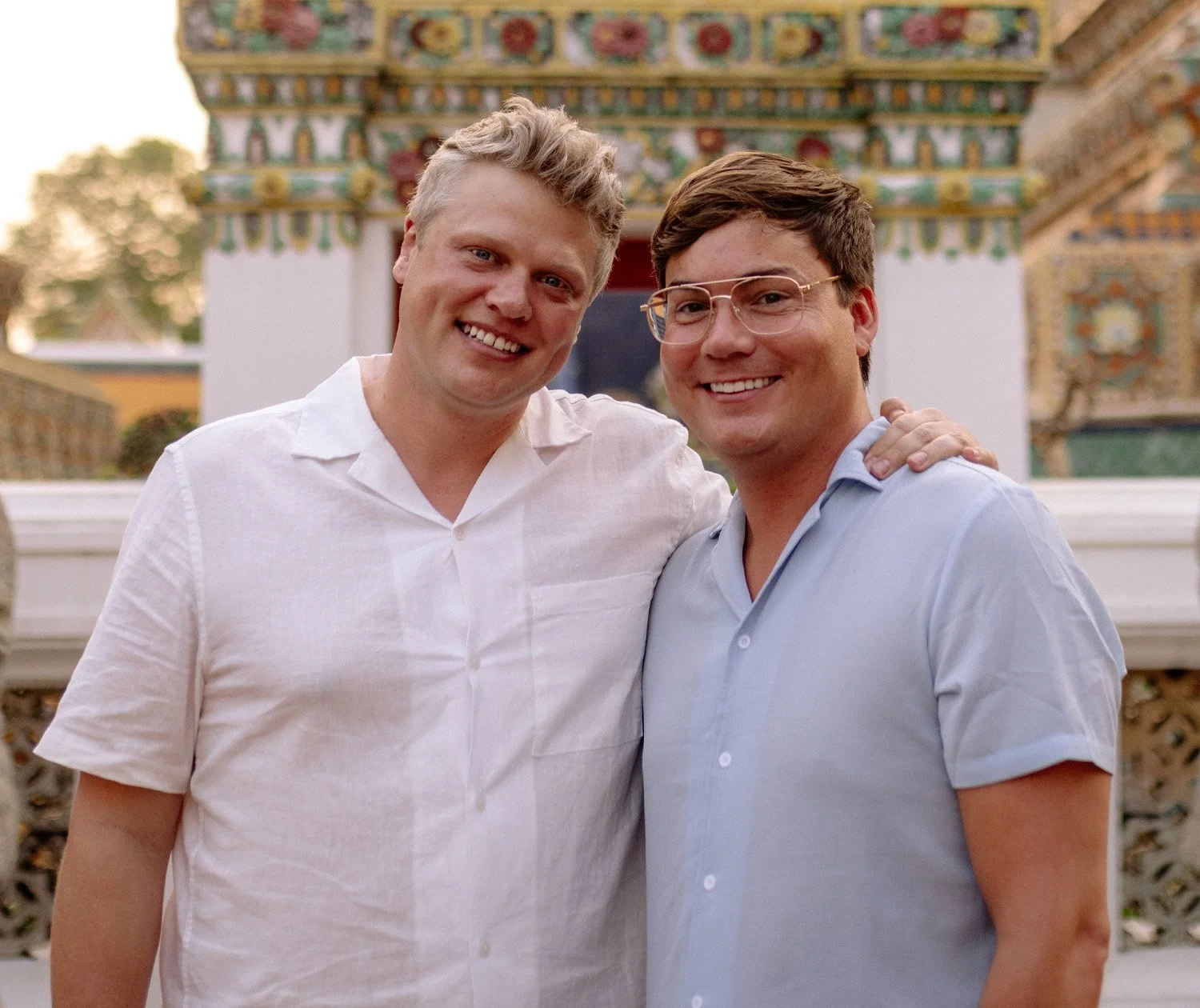 Two smiling men in light-colored shirts standing close together with one man's arm around the other's shoulder, in front of a colorful, ornate structure.