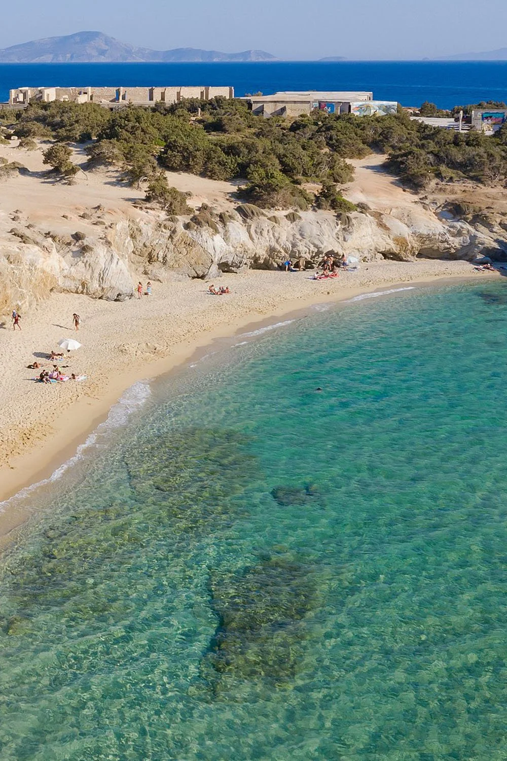 A sandy beach with clear turquoise water, people sunbathing and walking, rocky cliffs, green bushes, buildings in the background, and mountains across the water.