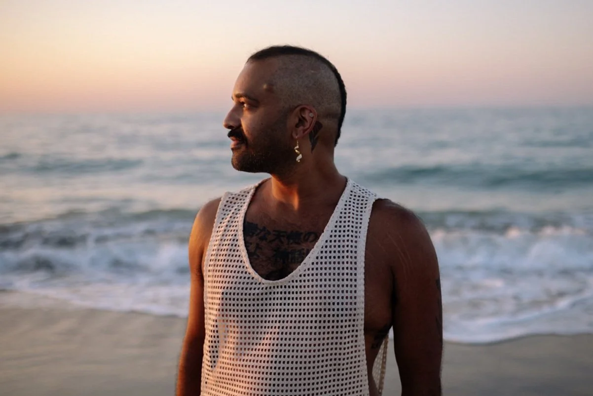 A man with tattoos, earrings, and a mohawk hairstyle standing on the beach during sunset, looking towards the ocean.