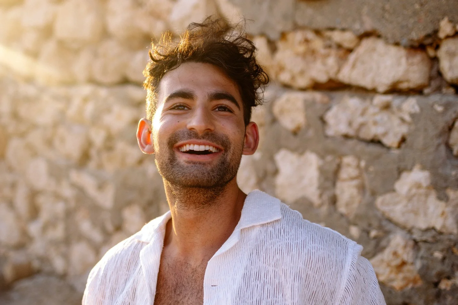 A smiling young man with dark, curly hair and a stubbled beard standing in front of a stone wall, wearing a white shirt, bathed in warm sunlight.