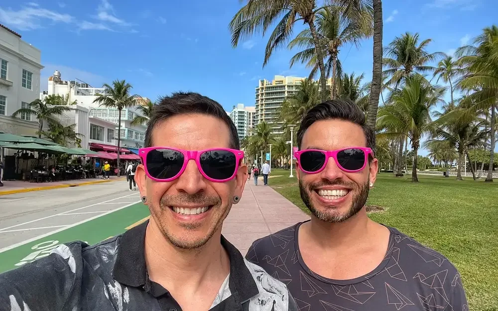 two men in pink sunglasses smile in front of palm trees and buildings