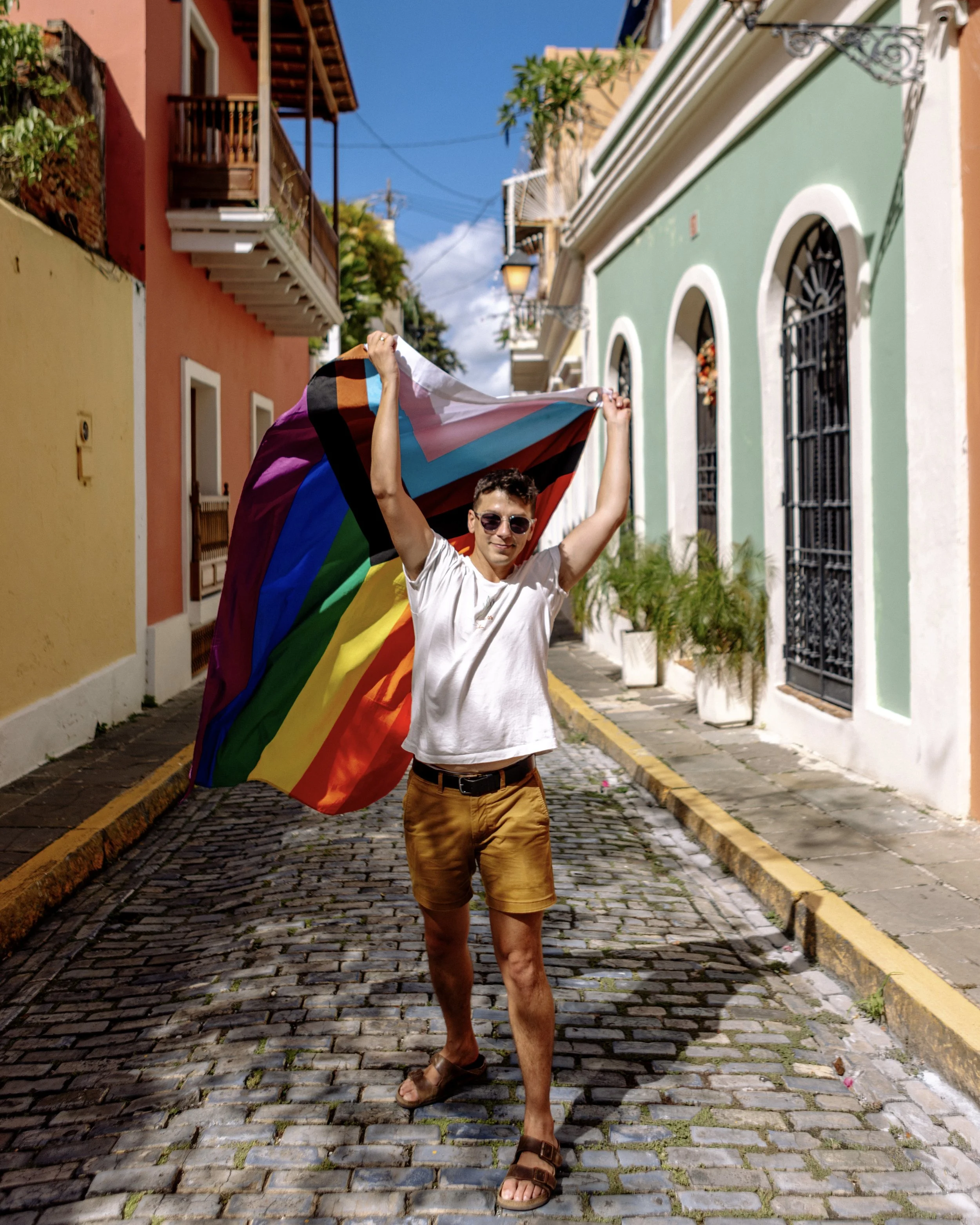 A person standing on a cobblestone street, holding a rainbow pride flag and smiling. colorful buildings line the street under a blue sky.