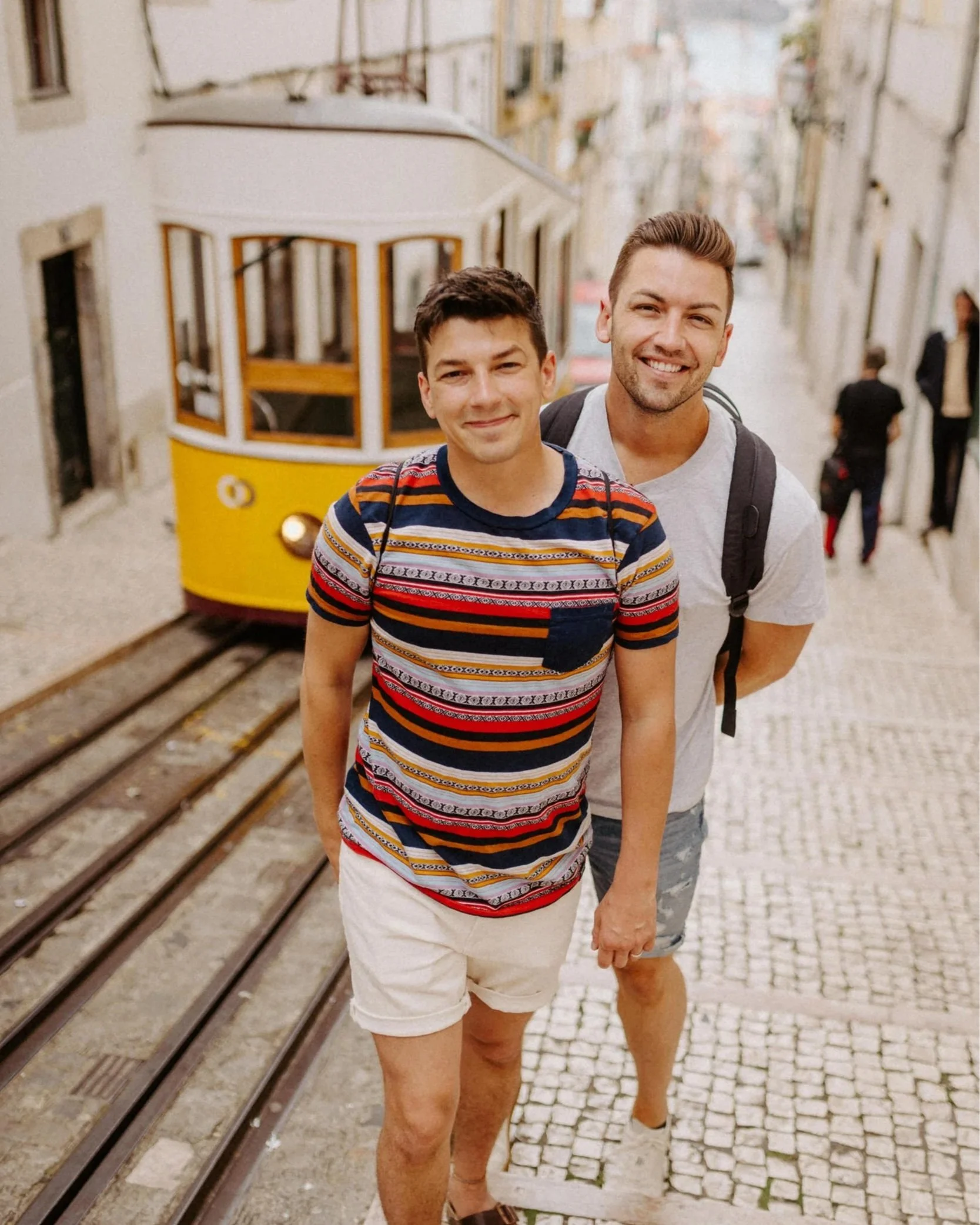 Two men smiling on a cobblestone hill in Lisbon, Portugal, with an iconic yellow tram behind them.