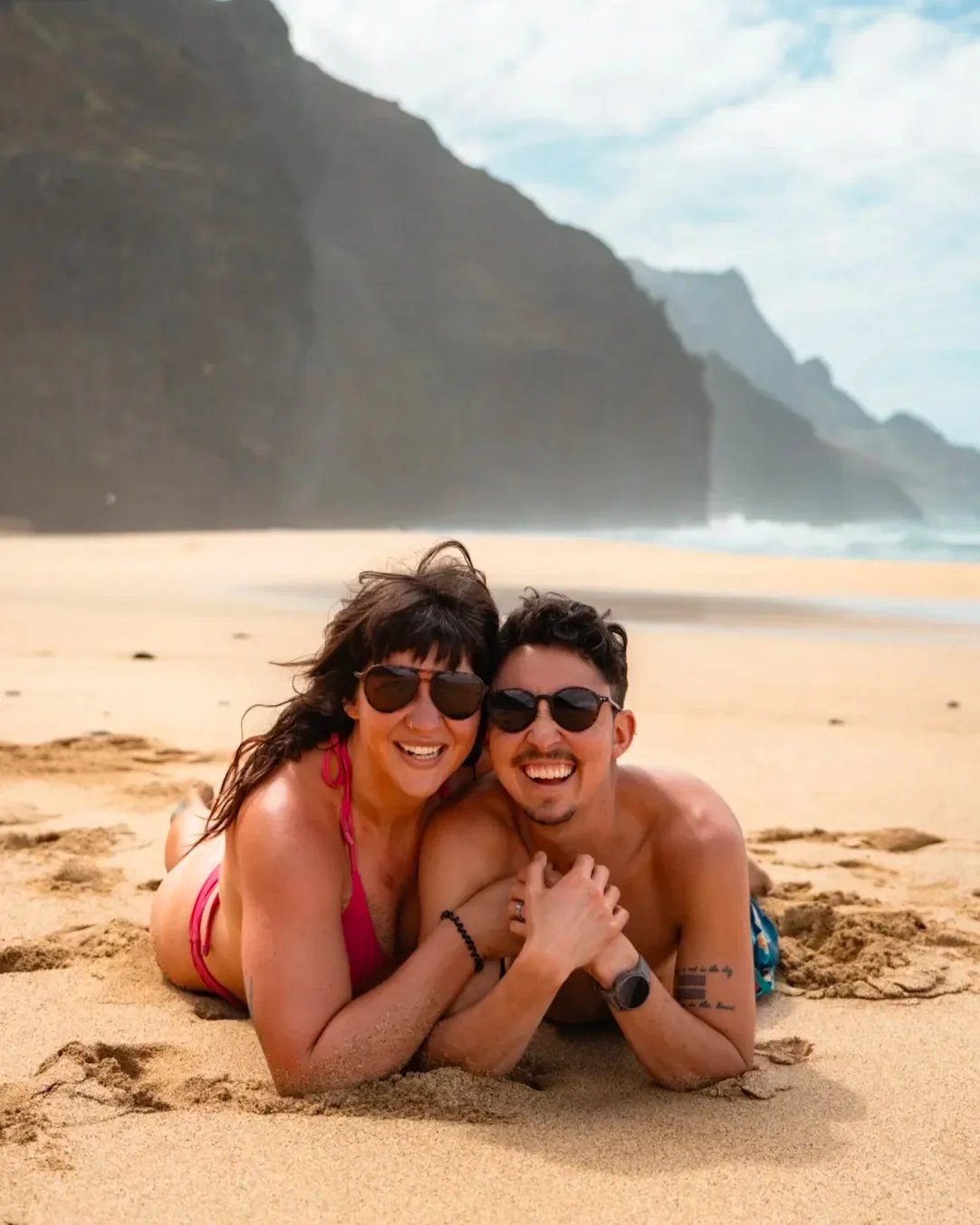 a queer couple embraces and smiles laying sand with ocean waves and cliffs in the background