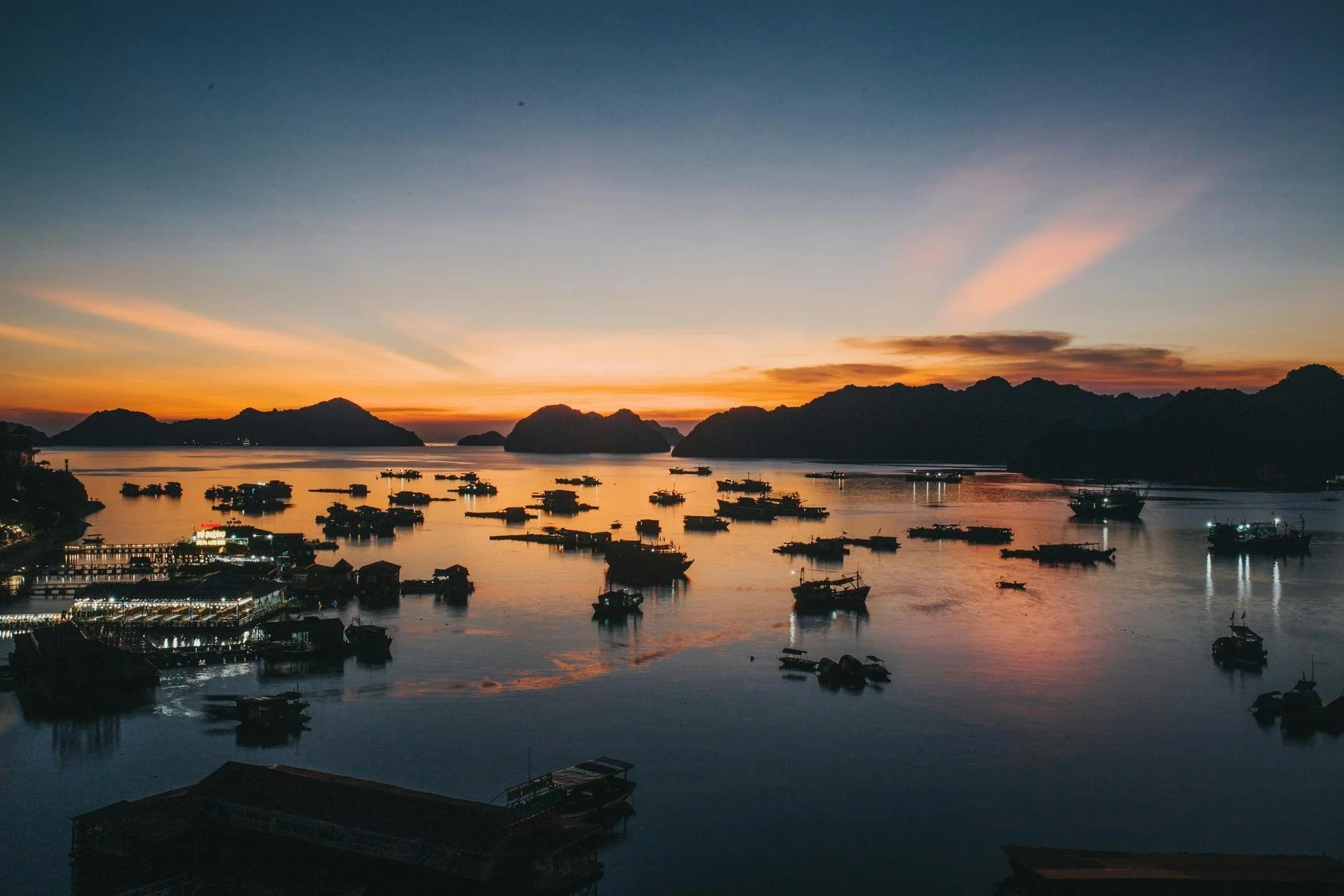 Dozens of fishing boats moored at sunset in Ha Long Bay, with karst mountain silhouettes and golden reflections.