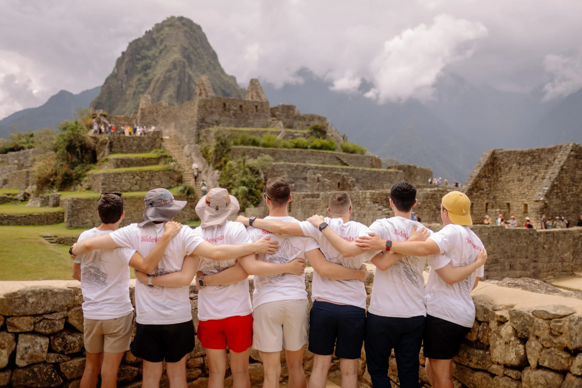 Seven friends in matching white SideQuests tees with arms around each other overlooking Machu Picchu ruins, Peru.