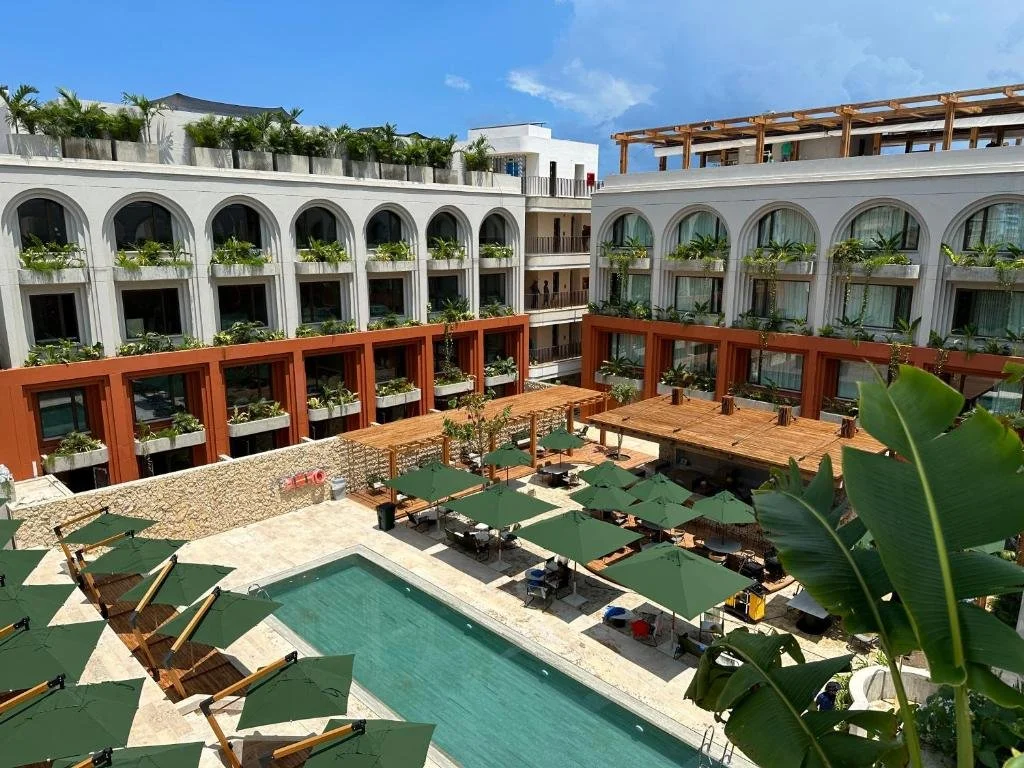 A hotel courtyard with a swimming pool, surrounded by lounge chairs with green umbrellas, and multi-story buildings with balconies and large windows.