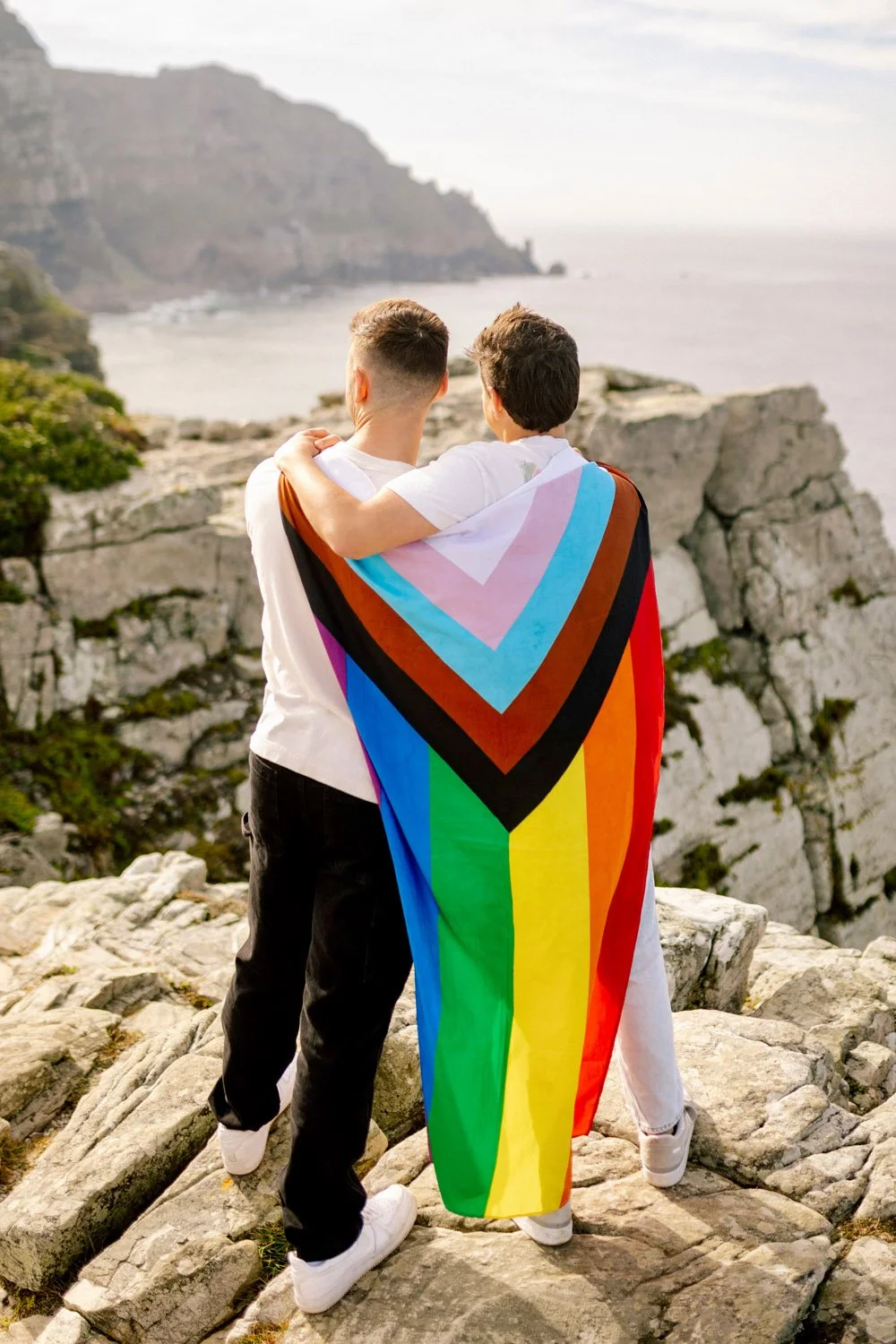 Two men standing on rocks overlooking the ocean, one wrapped in a rainbow pride flag, with cliffs in the background.