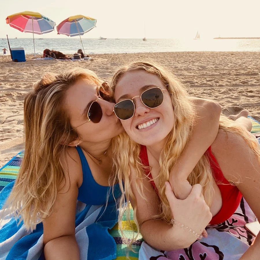 a lesbian blonde couple kisses and smiles while laying down on the beach and wearing sunglasses