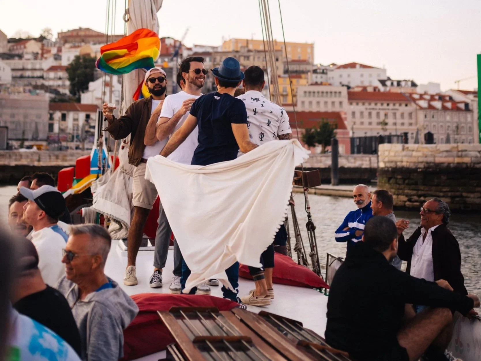 men dance and talk on a sailboat with a pride flag and Lisbon Portugal in the background