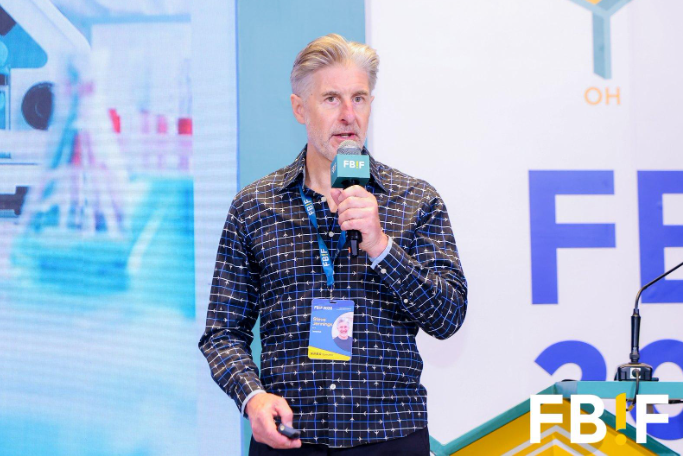 A man with gray hair and a beard speaking into a microphone at a conference, wearing a dark checkered shirt and a lanyard, with a large screen and conference branding in the background.