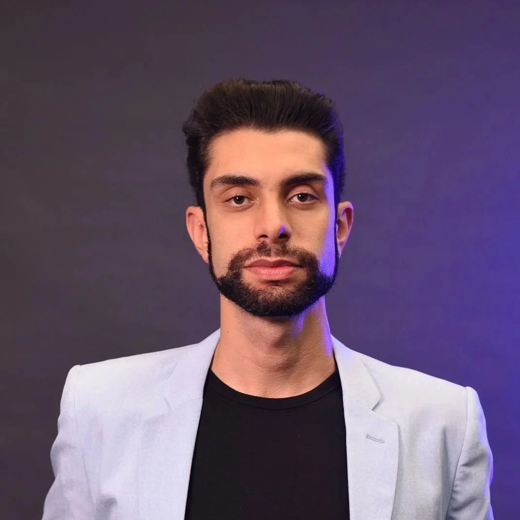 Portrait of a man with dark hair and a beard, wearing a light gray blazer over a black shirt, against a dark background with purple lighting.