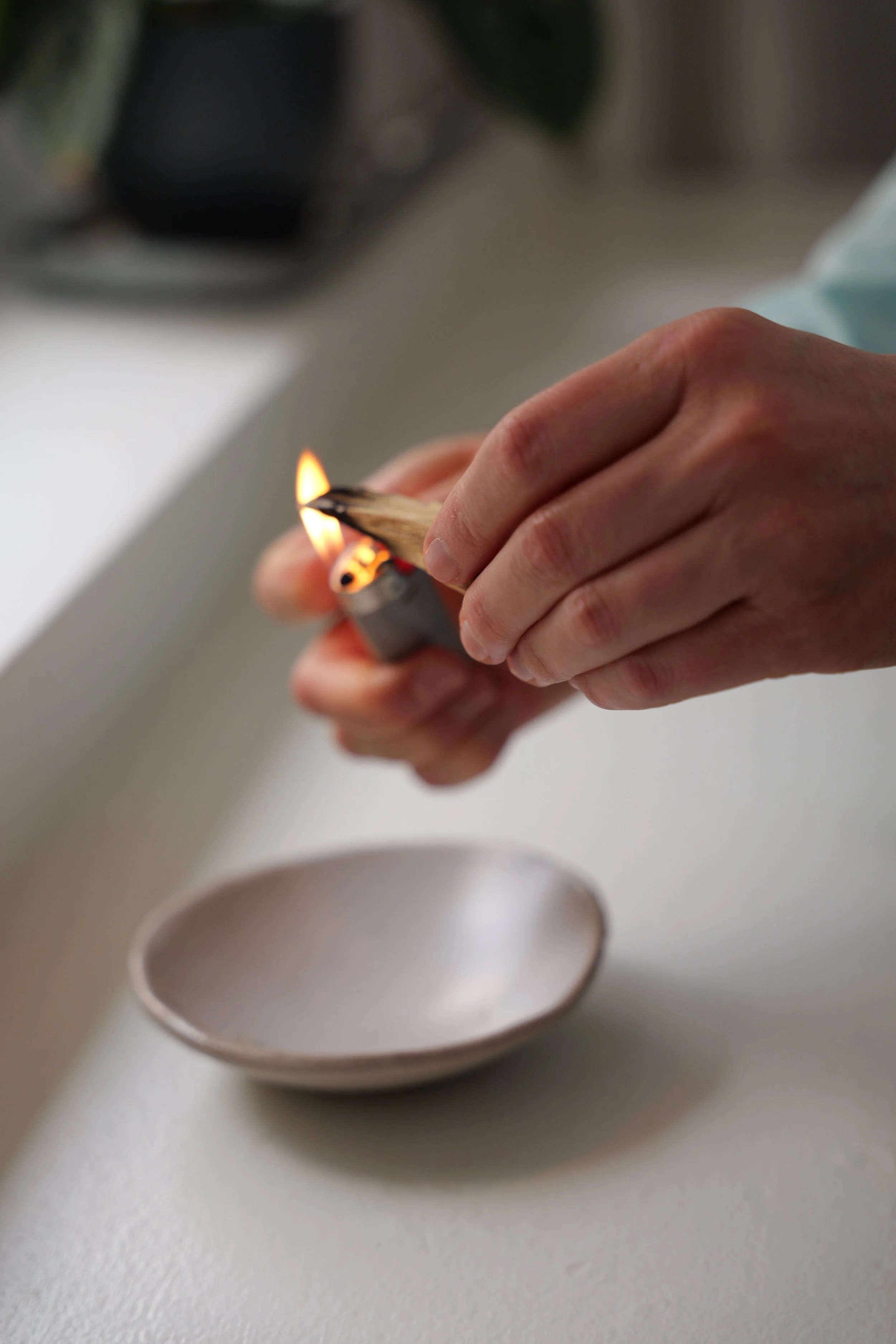 A person holding a lit matchstick over a small, empty ceramic bowl on a white surface.