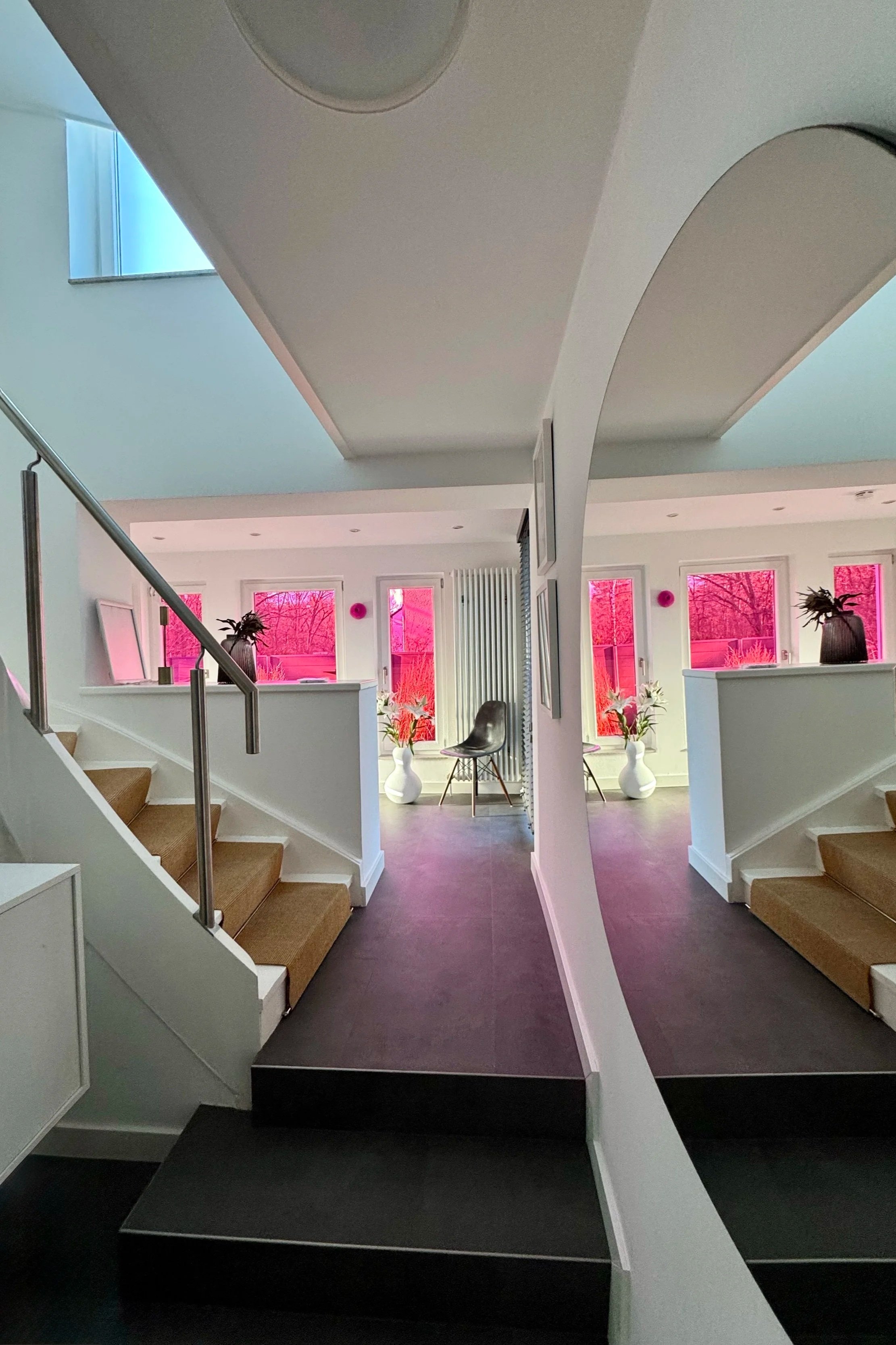 Innenansicht eines modernen Wohnraums mit Treppen, weißen Wänden, großen Fenstern mit pinken Sichtschutzvorhängen, Sitzgelegenheiten und Vasen mit Blumen.