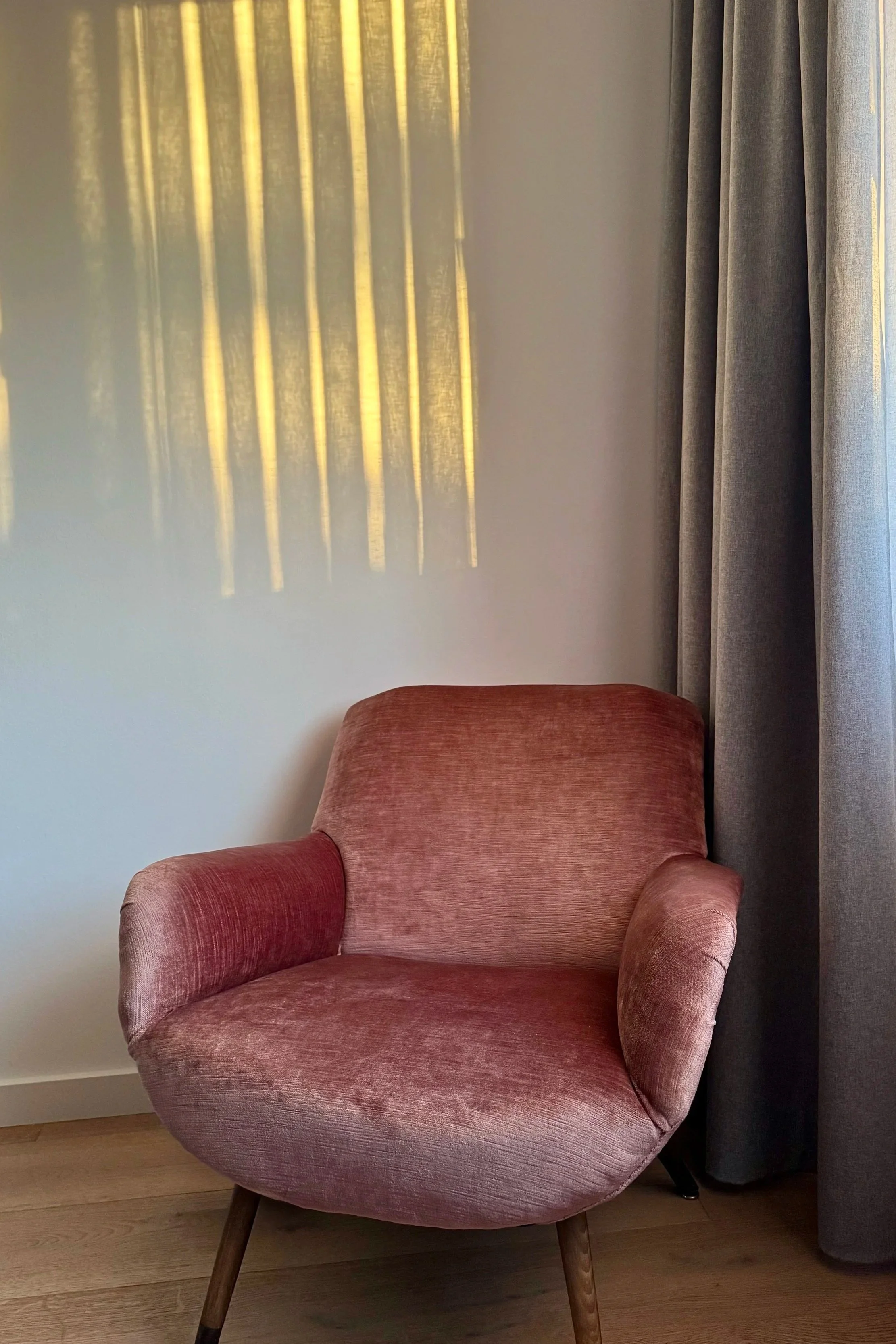 Ein rotes, gepolstertes Sessel in einem Raum mit hellen Wänden, grauen Vorhängen und Holzfußboden. Die Sonne scheint durch einen Fenster, was einen Schatten an die Wand wirft.