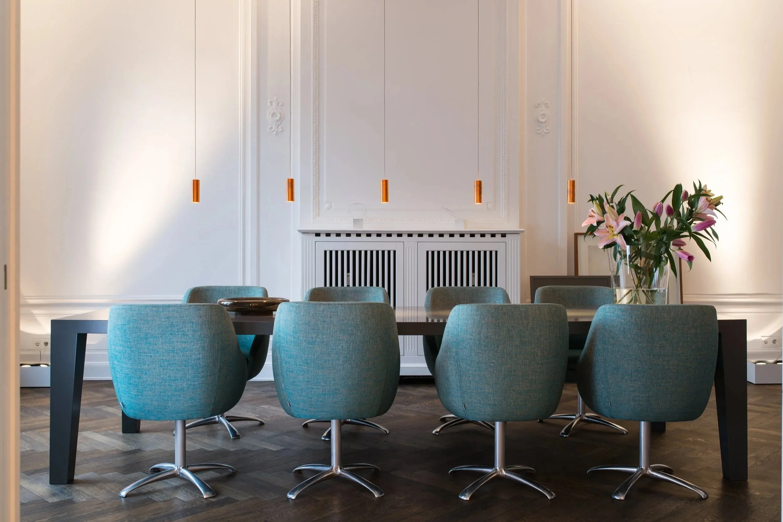 Ein moderner Konferenztisch mit acht blauen Sesseln in einem eleganten Raum mit weißen Wänden, dekorativer Wandgestaltung und einer großen Vase mit pinken Lilien.