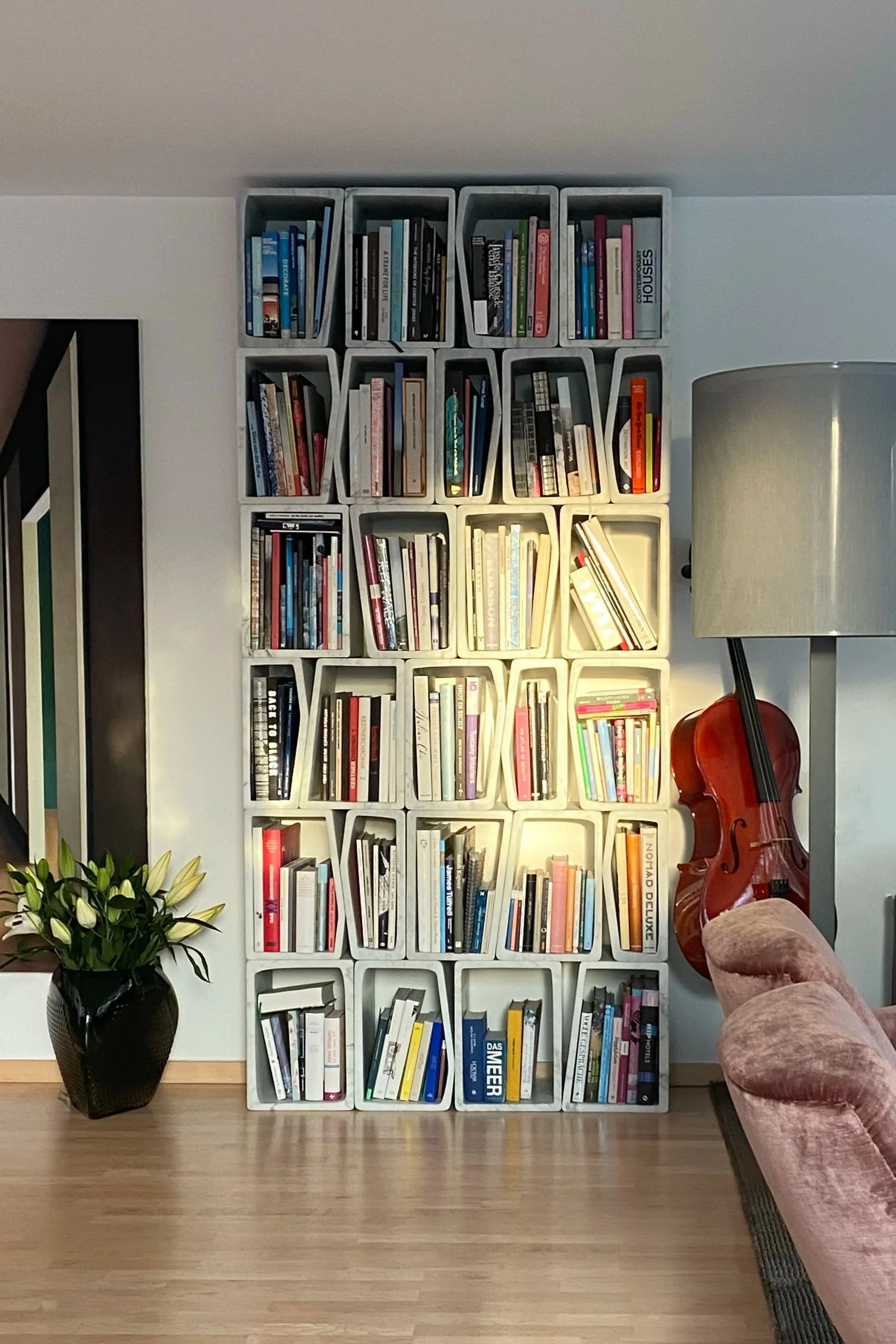 Ein Bücherregal mit mehreren Fächern, eine Stehlampe, ein Cello, eine pinkfarbene Couch und eine Vase mit weißen Blumen in einem Wohnzimmer.