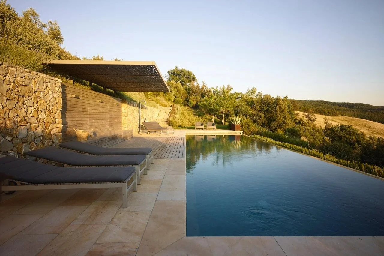 Ein Infinity-Pool im Außenbereich mit Liegestühlen und Sonnenliegen, umgeben von Bäumen und einer Landschaft mit Feldern unter klarem Himmel.