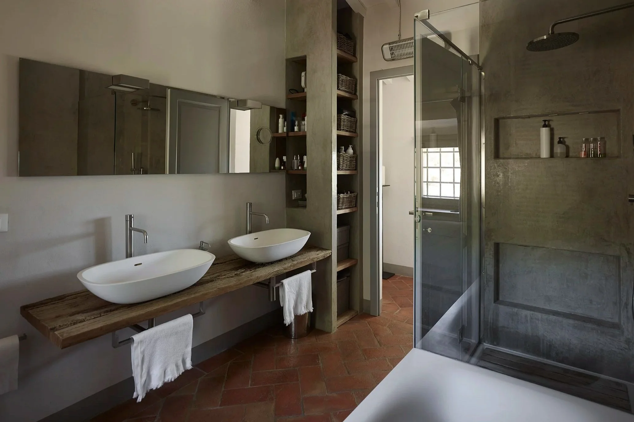 Ein modernes Badezimmer mit Doppelwaschbecken auf einem Holztisch, großen Spiegeln, offenen Regalen mit Körben, einer separaten Dusche mit Glaswand, braunen Fliesen und einem Fenster mit Raster.