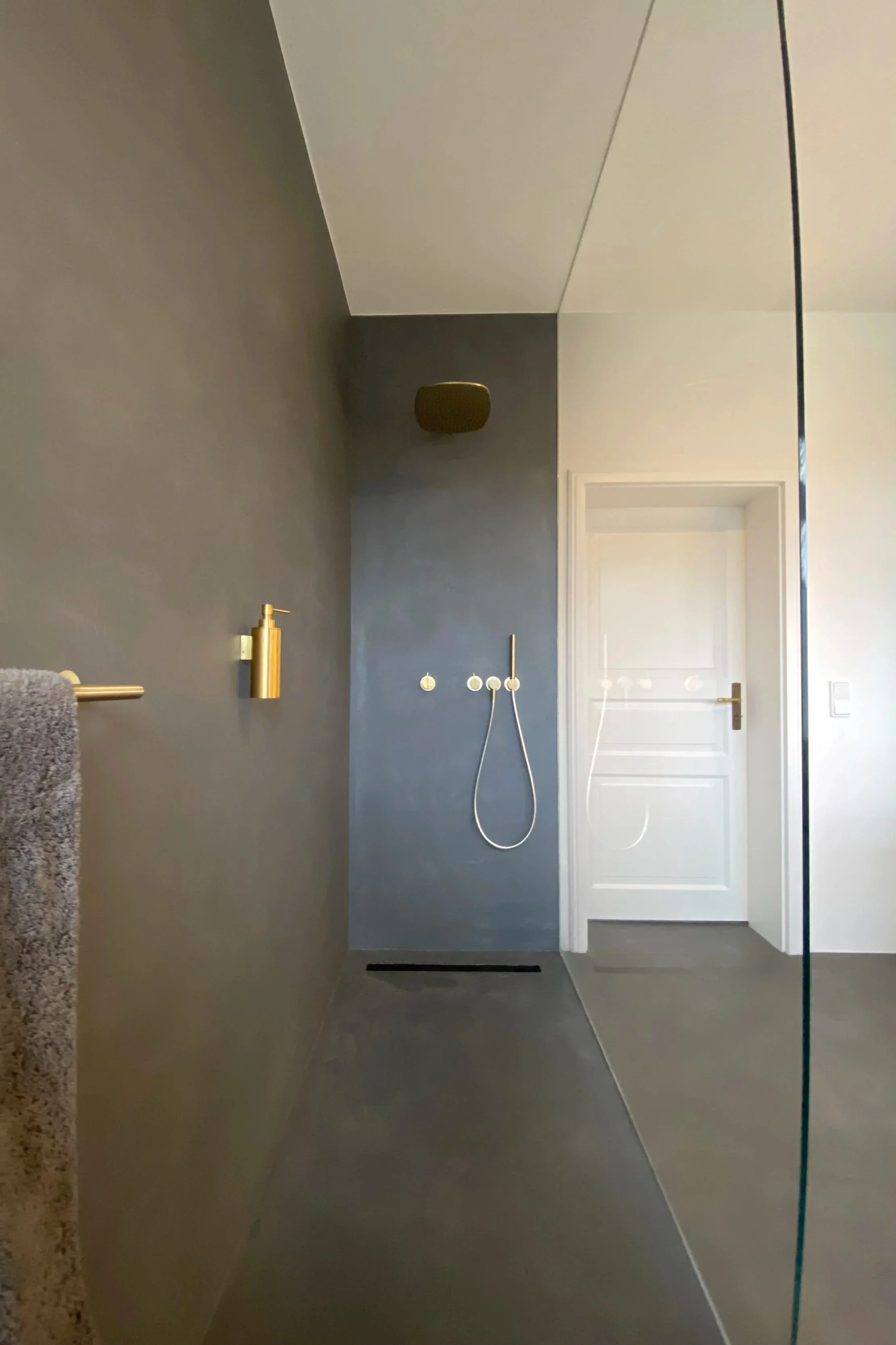 Moderner Badezimmerbereich mit dunkler Wand, Duschkopf, goldener Seifenspender, Handtuchhalter, Glaswand, Weißes Tür, grauer Boden, Wandleuchte